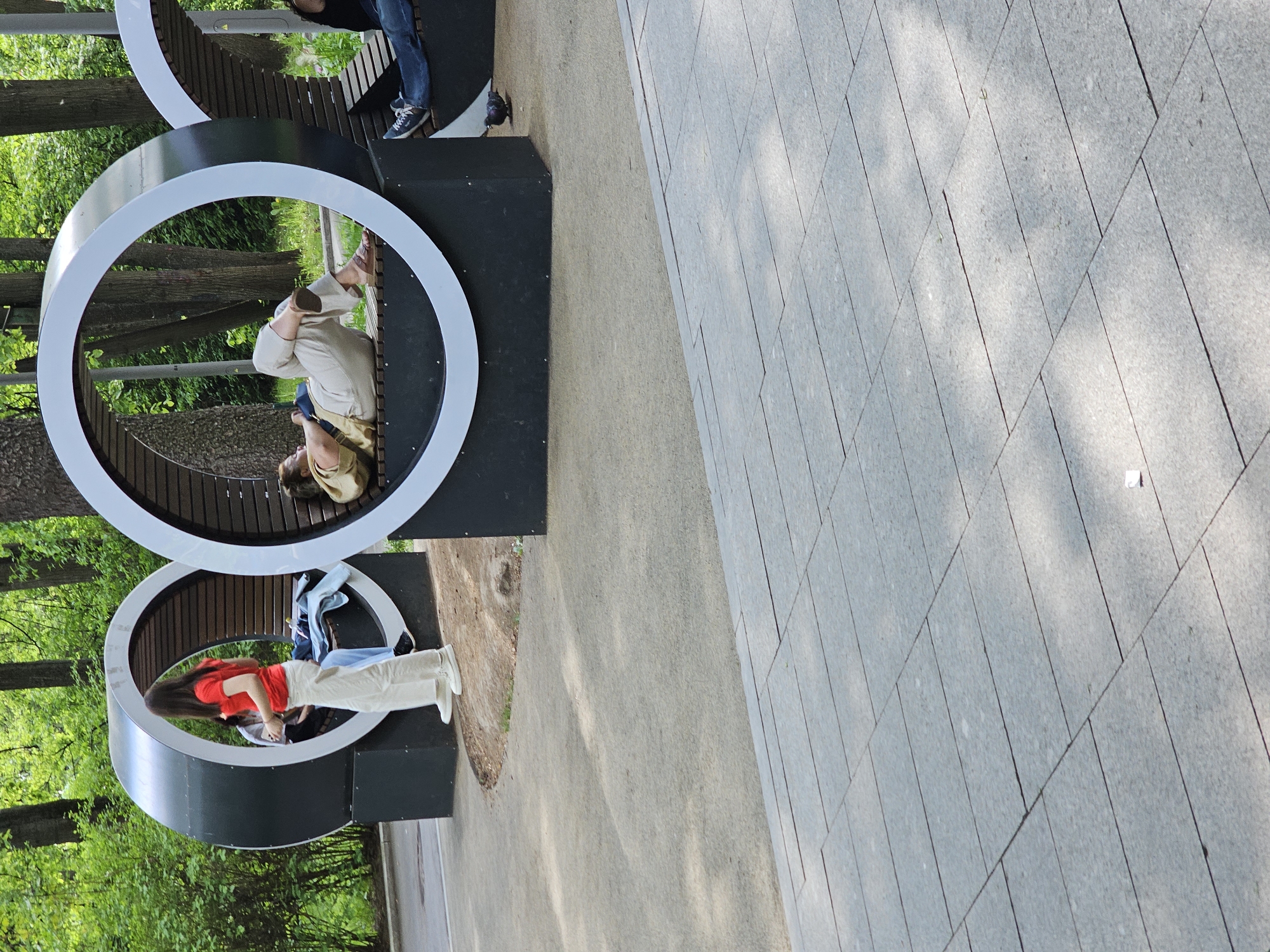 The image captures an outdoor scene in Moscow, Russia. Two people are seated on modern, circular-shaped benches with wooden slats within the circles. The benches are arranged in a stacked configuration. One person, wearing a red shirt and white pants, is sitting on the lower bench. The second person, in beige attire, is on the upper bench. The area surrounding the benches includes greenery, suggesting a park setting. A paved walkway borders the park area.
