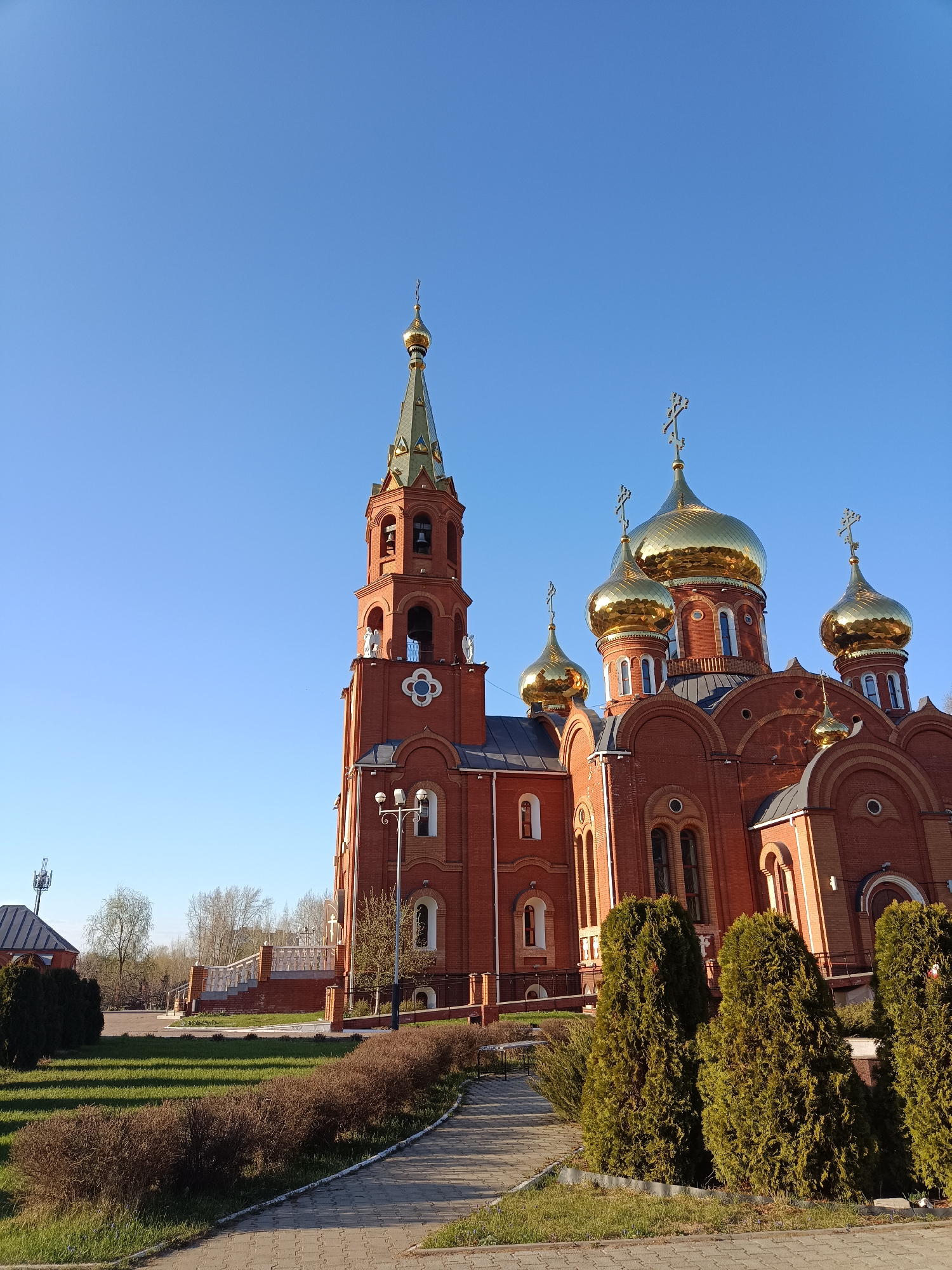 The image showcases a Russian Orthodox church located in the city of Unknown, Unknown. The church is constructed of red brick and features multiple golden domes topped with Orthodox crosses. A tall bell tower, also made of red brick, is located to the left of the main church structure. The tower is topped with a green spire and a golden dome. The sky is a clear blue. The church is surrounded by well-maintained green lawns, bushes, and a paved walkway. A lamp post stands next to the walkway. In the background, there are trees and a smaller building.