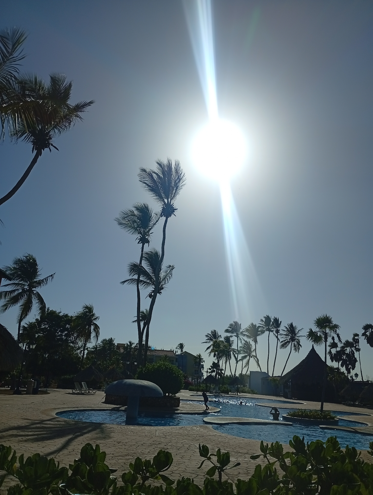 The image captures a sunny outdoor scene in Playa El Agua, Venezuela. A swimming pool with multiple sections and rounded edges is a central feature. The pool includes a mushroom-shaped structure in one section. Several people are visible in the pool, some standing on the shallow areas. Palm trees surround the pool area, casting shadows on the surrounding pavement. There are thatched-roof structures nearby. The sun is high in the sky, causing a bright glare and light rays. In the foreground, there are green plants. In the background, there are buildings with blue accents.