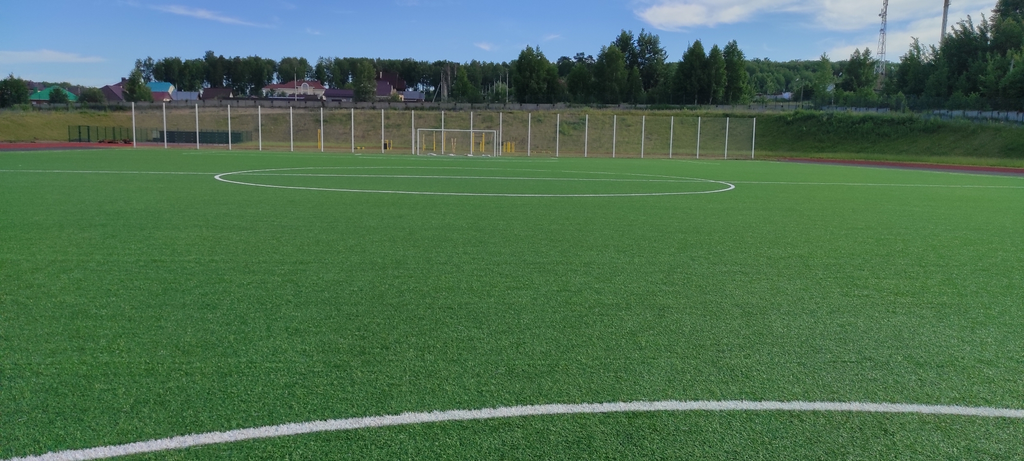 The image depicts a green artificial turf football field in Noviy, Russia, under a clear blue sky with scattered clouds. The field is marked with white lines indicating the center circle and halfway line. In the background, a chain-link fence separates the field from a grassy embankment, behind which are several houses with pitched roofs and trees. Further in the distance, more trees form a dense forest. The field appears to be part of a sports complex, with a running track visible along the left edge of the frame. Two tall towers, possibly communication masts, are visible on the right side of the horizon. There are no people or animals visible in the image.