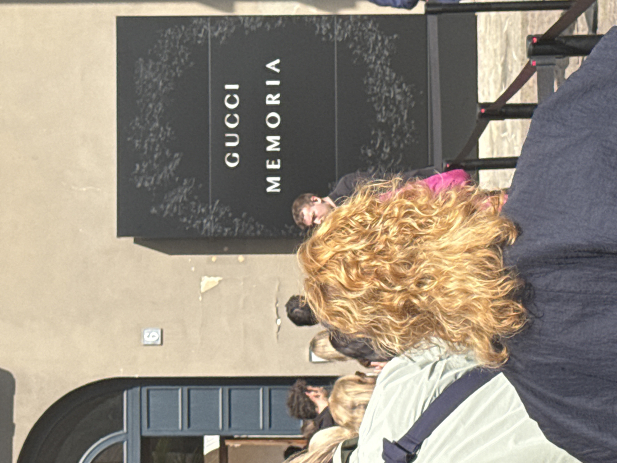 The image captures an outdoor scene in Milan, Italy, under bright daylight. Several people are visible, primarily from behind, appearing to queue or gather in front of a building. In the foreground, a person with prominent, curly blonde hair faces away from the viewer, wearing a light-colored top with a visible dark strap.
Behind this individual, other people with various hair colors are present, with one man in a dark shirt and a hint of pink clothing visible closer to the main sign. Partially visible on the right are barriers or stanchions, suggesting a structured waiting area or queue.
The central feature of the scene is a large, black sign affixed to the building's facade, displaying the white text "GUCCI MEMORIA." The building itself has a light beige or tan stucco finish. Towards the lower left, an arched entryway and a window or door with blue frames are discernible. A small square plaque with the number '6' is visible on the wall above the archway. The clear lighting indicates a sunny day. The overall activity suggests people are waiting to enter a Gucci-related exhibition or venue.