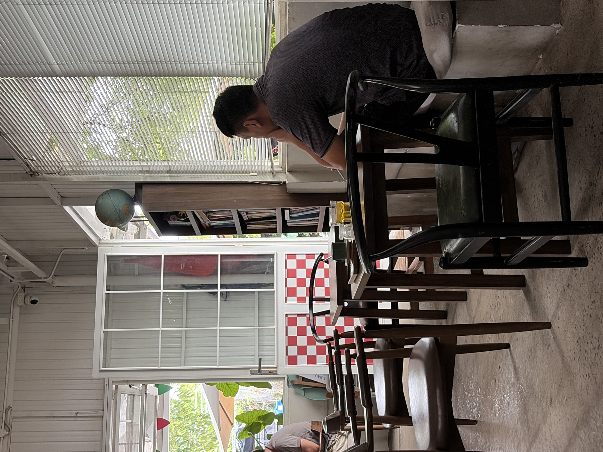 The image depicts the interior of a cafe in Da Nang, Vietnam, characterized by a rustic and eclectic design. A man, viewed from the back, is seated at a table near a window covered by blinds, appearing to be contemplating or waiting. He is wearing a dark patterned short-sleeved shirt. In the foreground, several wooden chairs with leather seats are arranged around tables, with one dark-colored chair prominently in the right foreground. A glass of amber liquid, possibly tea or coffee, sits on a nearby table.
To the left, an open door reveals an outdoor area with greenery and a partially visible umbrella, suggesting a connection to the city's natural surroundings. A red and white checkered pattern adorns the lower part of the door frame. A bookshelf filled with books stands against the wall, topped with a globe, adding to the cafe's intellectual ambiance. The cafe's architecture features corrugated metal walls and ceiling, with exposed pipes and a security camera visible. The flooring is a speckled concrete. The overall impression is of a relaxed, perhaps bohemian, establishment within Da Nang.