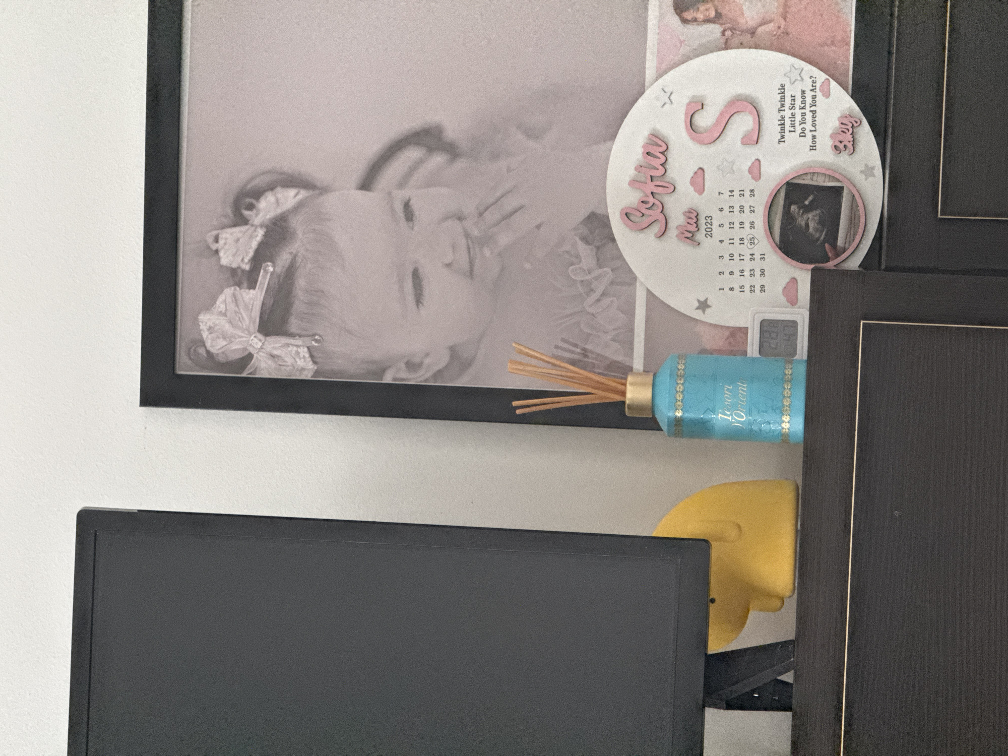 A framed black and white photograph of a baby girl with bows in her hair is displayed. To the right of the photograph is a circular wooden decoration with the name "Sofia" and the month "May 2023" printed on it. There is also a calendar section, a picture of a fetus, and the words "Twinkle Twinkle Little Star Do You Know How Loved You Are?". Next to this decoration is a teal diffuser bottle with a gold cap and reeds. A yellow duck-shaped object is on the left side of the image, in front of a black monitor. The scene is set indoors, with a white wall in the background and a dark wooden surface in the foreground. The image does not provide any information about its location in Chișinău, Moldova.