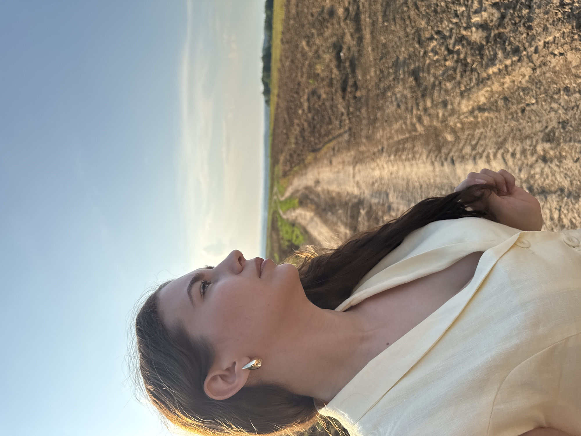 A young woman with long, dark hair stands outdoors in a field, facing to the right of the frame, looking into the distance. She is wearing a light yellow blazer and a gold earring. The background shows a dirt path winding through a field with some green patches and dry soil. In the far distance, a line of trees is visible under a clear blue sky. The lighting suggests it is late afternoon. The location is identified as Lukintsy, Russia.