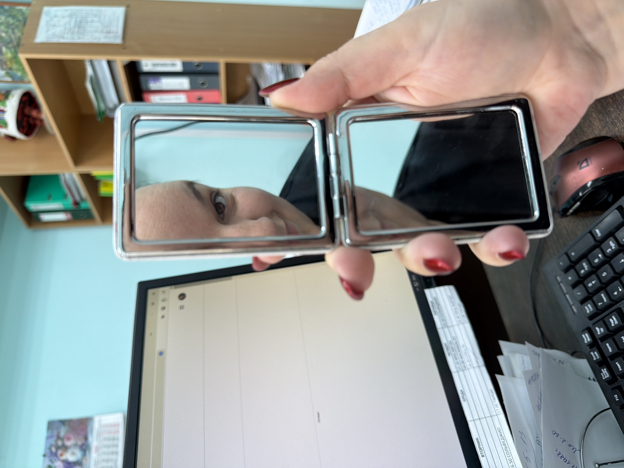 An overhead view, rotated 90 degrees counter-clockwise, captures a scene within an office or workspace in Chaikovsky, Russia. The central focus is a person's hand, adorned with red nail polish, holding an open, silver-framed compact mirror. The left panel of the mirror reflects a close-up of the person's face, showing an eye, eyebrow, and part of the cheek, indicating they are looking at their reflection.
Behind the mirror and hand, a light blue wall forms the background, above which a wooden shelf unit is visible, filled with binders in various colors, including red, black, and green. Below the mirror, on a desk, a computer monitor displays a light, mostly blank screen. To the right of the monitor, a black keyboard and a metallic copper-colored computer mouse are positioned. Several documents and papers are scattered on the desk, with some text partially legible.
Visible text from documents includes Russian words like "ВТОРНИК" (Tuesday) and "СРЕДА" (Wednesday), along with "6.50 СОЛЬФЕДЖИО" (6.50 Solfeggio) and "1720 ПАНА" (1720 Pana). Other numerical and alphanumeric sequences such as "5-15", "11.19881", and "Лен.2-60" are also present. Keyboard keys "F12", "Insert", "Home", "Page Down", "5", "9", "3", and "#" are clearly identifiable. The overall setting suggests a momentary pause from work, perhaps for a quick check in the mirror.