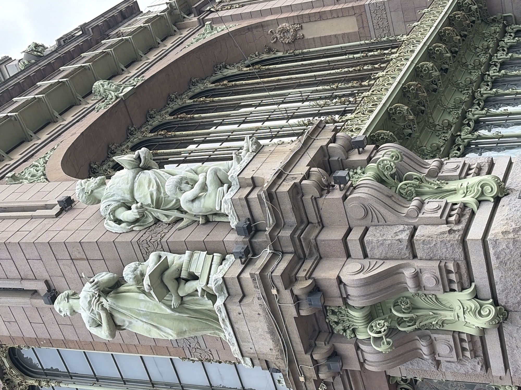 The image shows a low-angle view of the Singer House, also known as the Dom Knigi (House of Books), in Saint Petersburg, Russia. The building features a striking corner with sculpted figures and ornate architectural details. On the left, a light-colored statue depicts a woman holding a stylus and palette, with a young child reading a book at her feet, perched on a stack of books. To the right, another statue shows a figure, possibly male, holding a book. The building itself is constructed of reddish-brown granite blocks. Elaborate green metalwork, characteristic of Art Nouveau style, adorns the balconies and window frames. The windows are tall and arched, with multiple panes. The sky is overcast. The scene is static, with no visible people or animals. The lighting suggests it is daytime.
