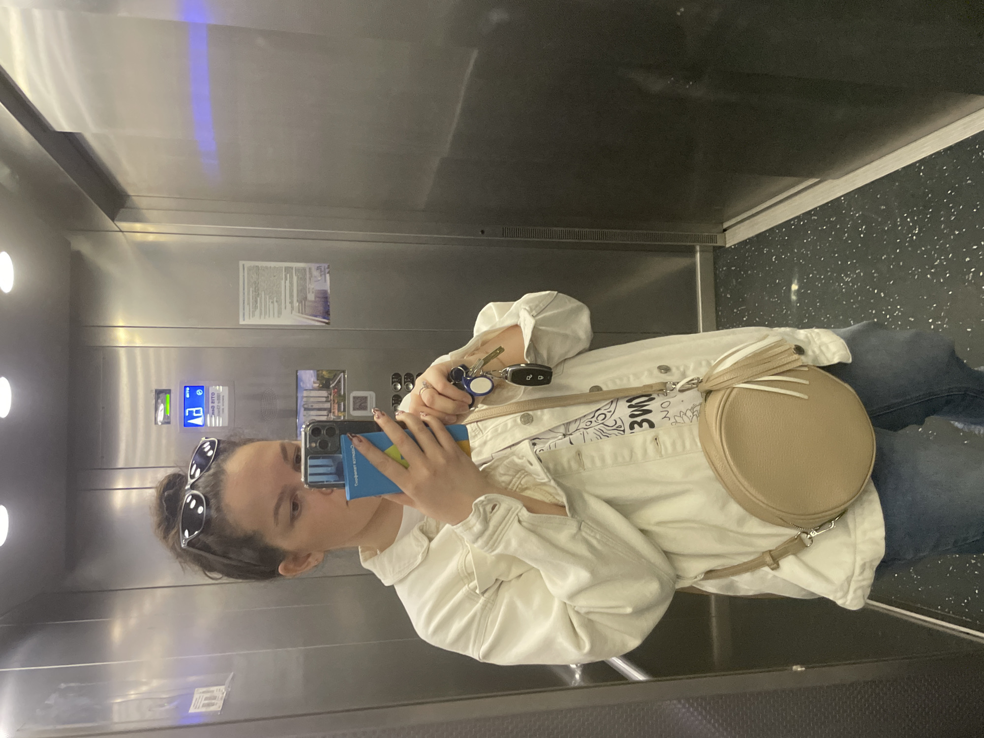 A young woman is taking a selfie in an elevator. She has her hair in a bun and sunglasses are atop her head. She is wearing a white jacket, jeans, and a crossbody bag. She is holding her phone in one hand and keys in the other. The elevator has stainless steel walls and a speckled floor. The interior includes a sign and control panel with buttons in Russian. The elevator's location can be identified as Izhevsk, Russia, based on a sign in Russian mentioning the city.