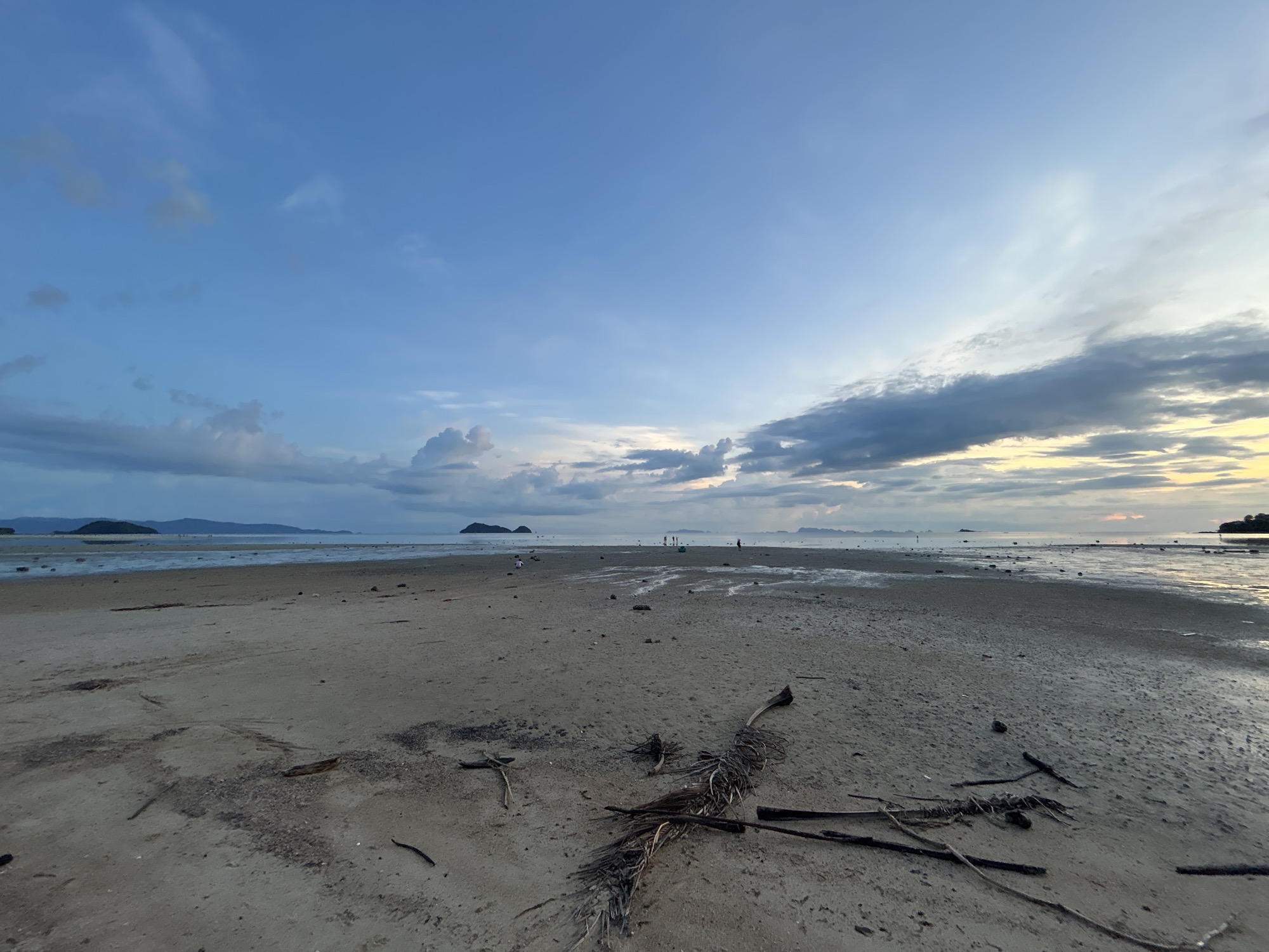 The image shows a wide, sandy beach with the ocean in the background. The tide is out, revealing wet sand and shallow pools of water. There are scattered pieces of driftwood and vegetation on the sand. In the distance, there are several small islands and the silhouettes of landforms. The sky is a pale blue with scattered clouds, and the sun appears to be low on the horizon, suggesting either sunrise or sunset. There are a few small figures of people walking on the beach in the distance, some of whom appear to be children. The overall scene is tranquil and natural. The location is identified as Koh Phangan, Thailand.