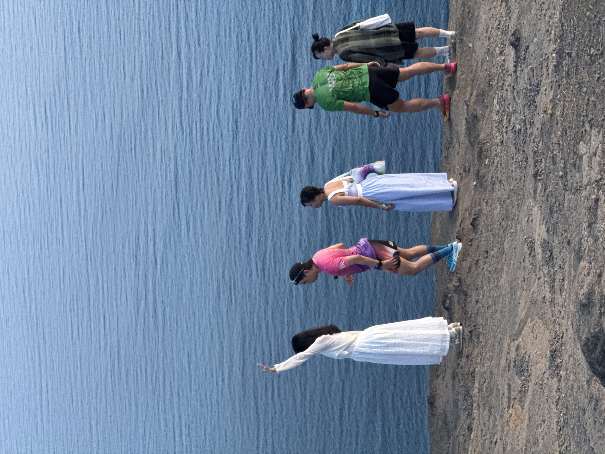 Five people are standing on a rocky cliff overlooking a large body of water. The water appears calm with small ripples. The group is positioned from left to right: a woman in a long white dress with her left arm raised, a person in a pink and purple tie-dye shirt and black shorts, a woman in a long light blue dress carrying a bag over her left shoulder, a man in a green t-shirt and black shorts, and a person in a dark checkered jacket and black shorts. The cliff is covered in sparse vegetation and rocks. The sky is not visible in the frame. The location is the coast of Lý Sơn Island, Quảng Ngãi Province, Vietnam.