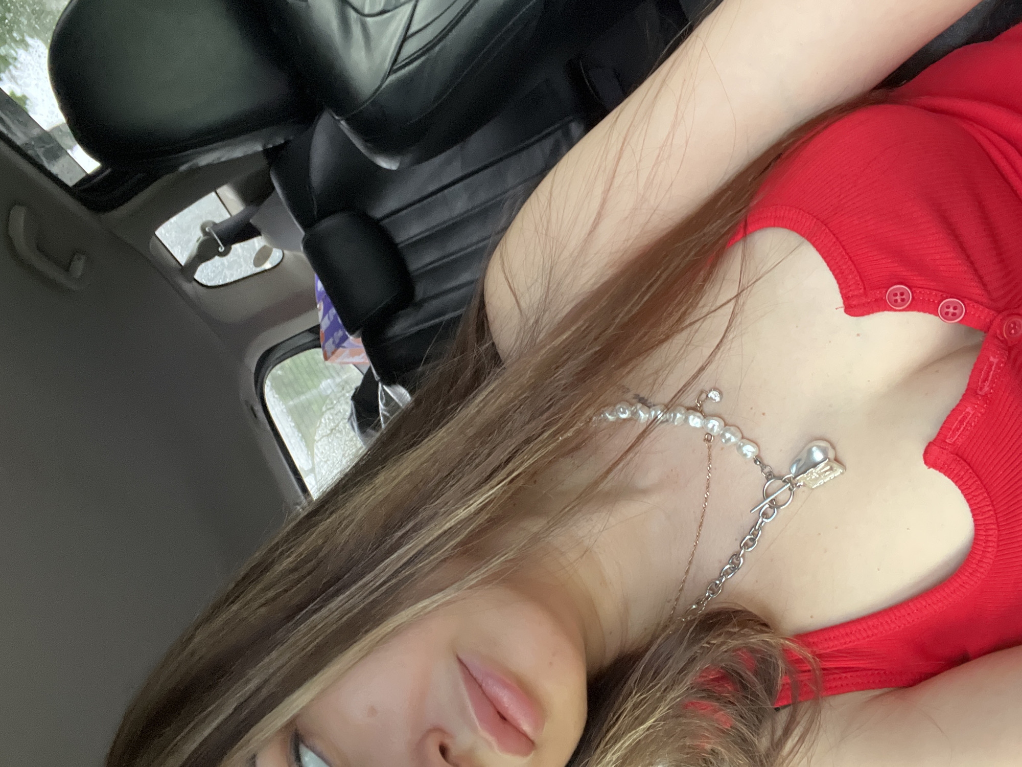 The image shows a close-up, diagonal view of a person inside a car, likely taking a selfie. The subject is a young woman with long, light brown hair featuring lighter highlights, partially obscuring her face. Her lips are visible, appearing full and pink, along with a hint of her chin and one eye.
She is wearing a red, ribbed top or dress with small red buttons visible along the side of the garment. Around her neck, she has two layered necklaces: one is a string of white pearls, and the other is a silver chain with a heart-shaped pendant and a toggle clasp closure.
The background reveals parts of the car's interior. A black leather seat is partially visible behind her. Above, a rearview mirror and an open sunroof are seen, with light gray, cloudy or rainy weather visible through the sunroof and a side window. A small blue and purple item, possibly an air freshener, hangs near the rearview mirror. The overall lighting suggests it is daytime, but overcast. No text is visible in the image.
