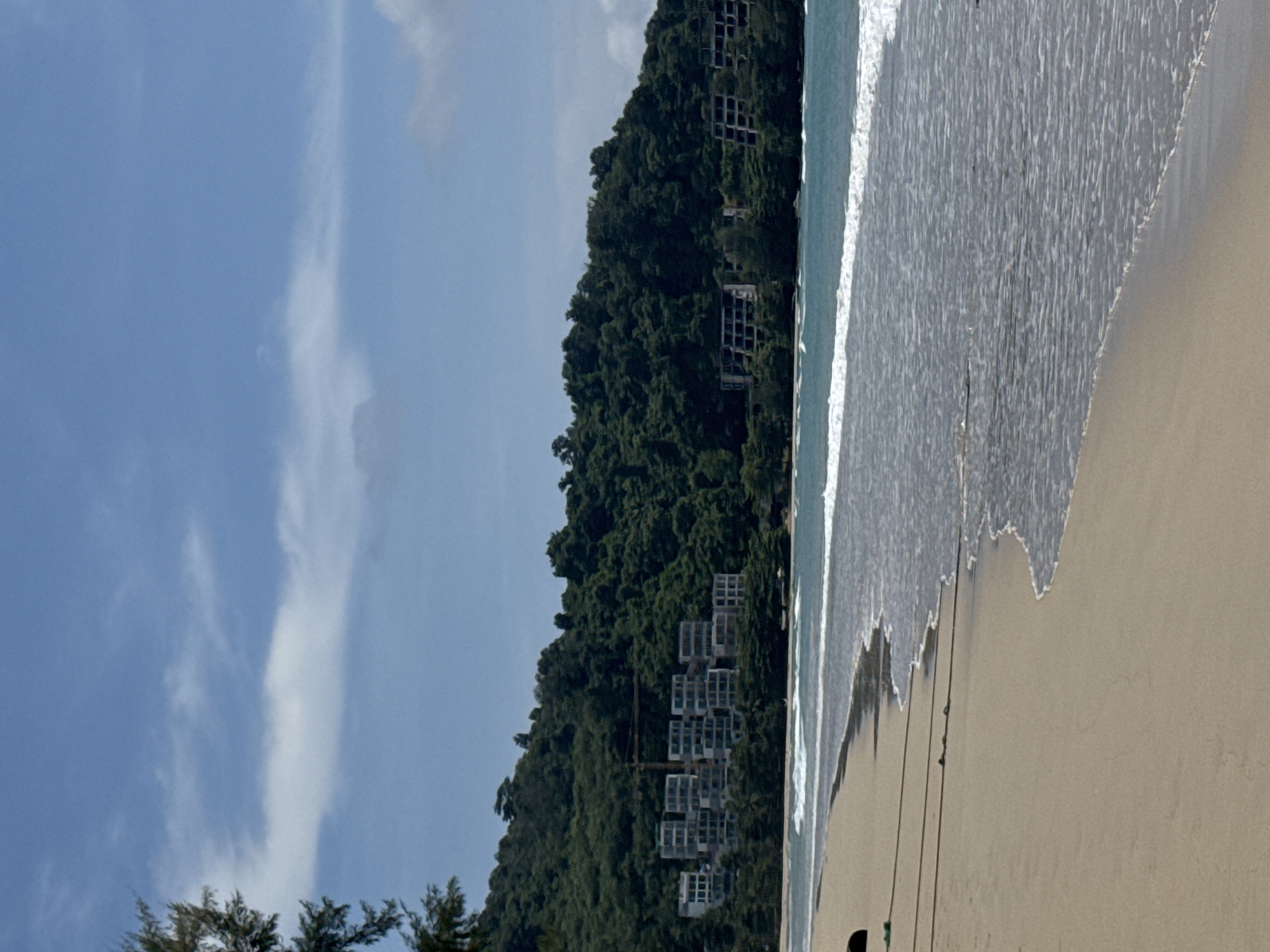 The image depicts a tropical beach scene in Sakhu, Thailand, featuring a sandy shore, gentle ocean waves, and a densely forested hillside in the background. Several unfinished or abandoned multi-story buildings are visible on the hillside, suggesting a construction project in progress or halted. The sky is partly cloudy with a clear blue expanse. A tree branch extends into the frame from the upper left, and two thin lines, possibly ropes, stretch across the sand in the foreground. The overall impression is one of a developing or partially developed coastal area.