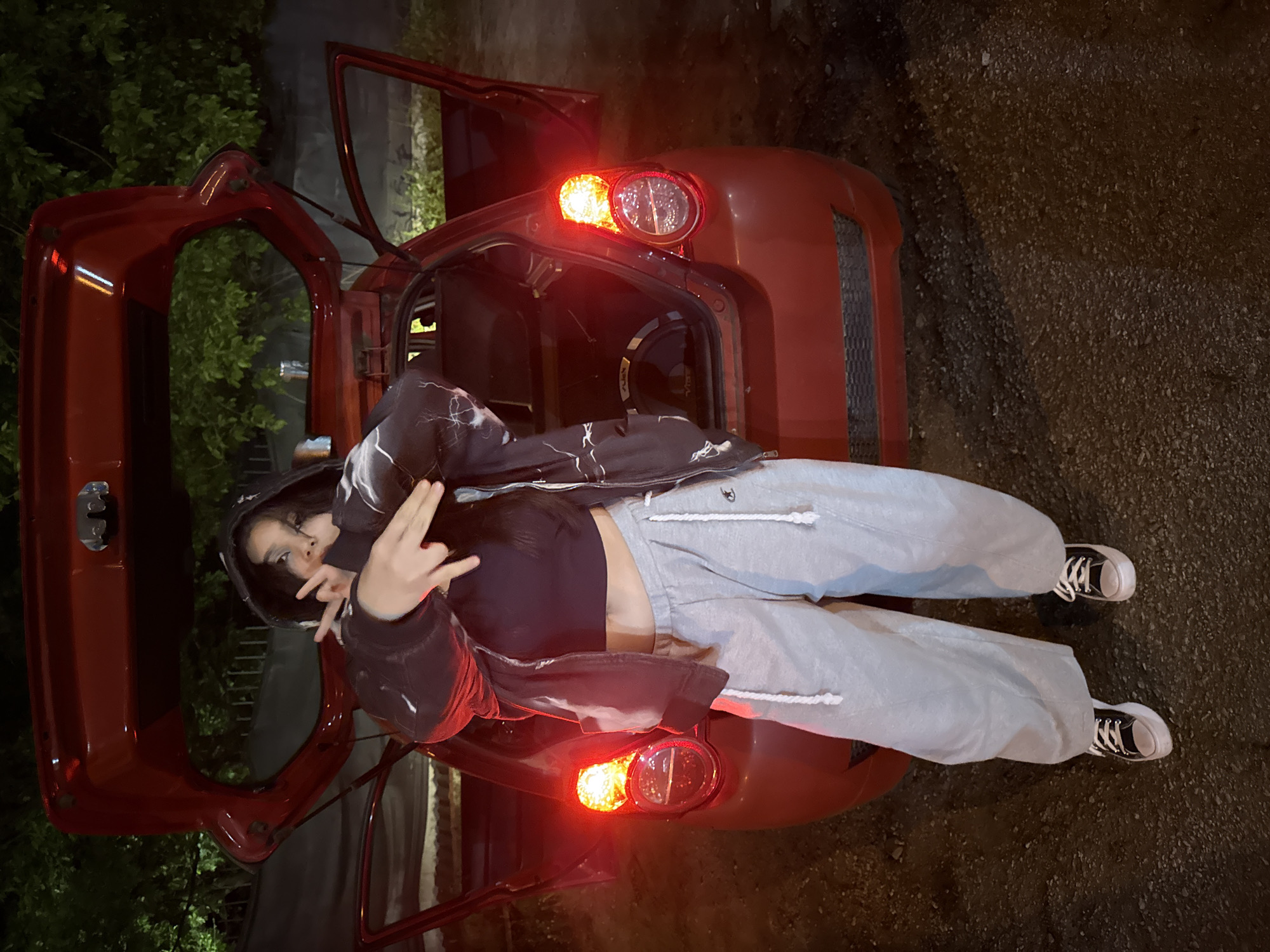 A young person with long dark hair, wearing a black cropped top, grey sweatpants, and a black hooded jacket with a white pattern, is lying across the open trunk of a red compact car. The car's taillights are illuminated. The person is making a hand gesture with both hands held up near their face. The car's doors are open on both sides. The surrounding environment appears to be a dimly lit outdoor area with trees and pavement. The exact location in Blagoevgrad, Bulgaria is not possible to determine.