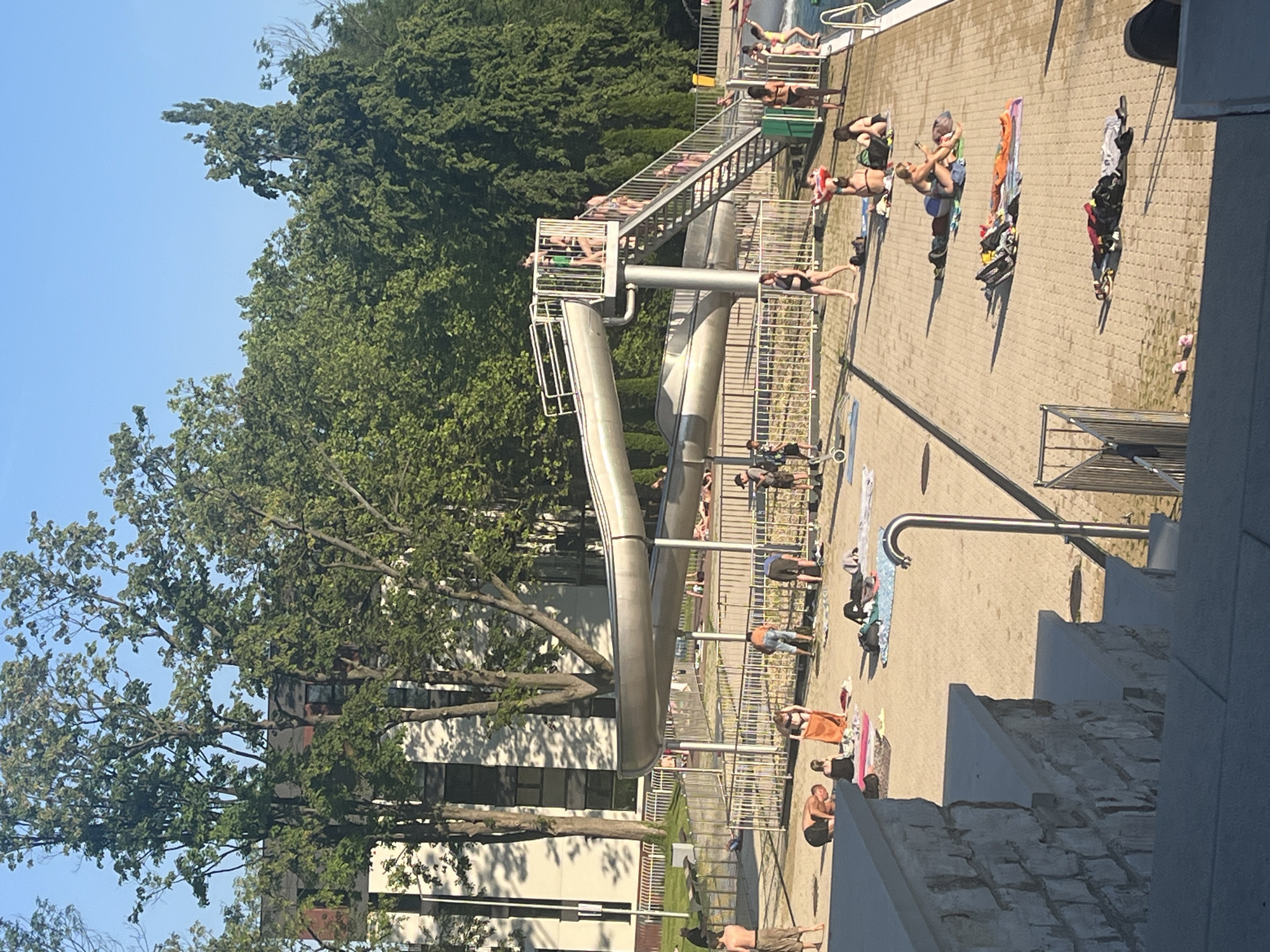 The image shows an outdoor water park on a sunny day. A large metal water slide structure dominates the center of the frame, with people visible on the stairs and at the top. The ground is paved with tan brick pavers. Several people are sunbathing or relaxing on towels and yoga mats scattered across the paved area. Some individuals are standing, others are sitting or lying down. A group of people are gathered near the base of the slide structure, with some appearing to be preparing to go down or just having exited. In the background, there are trees and a modern-looking building. A swimming pool is visible on the right side of the image, with a few people in or near the water. The scene depicts a recreational environment with various activities taking place.
