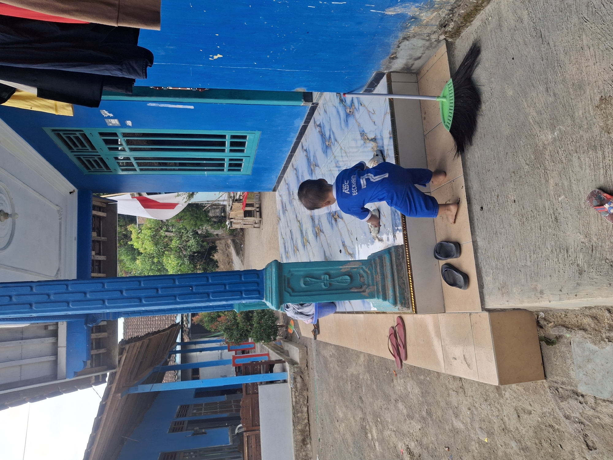 The image depicts a young child in a blue shirt with the name "BECKHAM" and the number "7" on the back, wearing blue pants. The child is on a tiled porch, seemingly cleaning the floor with their hands. A green broom with black bristles lies on the concrete ground in front of the porch. Red flip-flops are placed on the tiled edge of the porch, and black flip-flops are on the steps. Behind the child, a blue house with a green-framed window is visible. Clothes are hanging on a line outside the house. The surroundings appear to be an outdoor area with dirt, plants, and other buildings in the background, suggestive of a residential setting in Muncar, Indonesia.