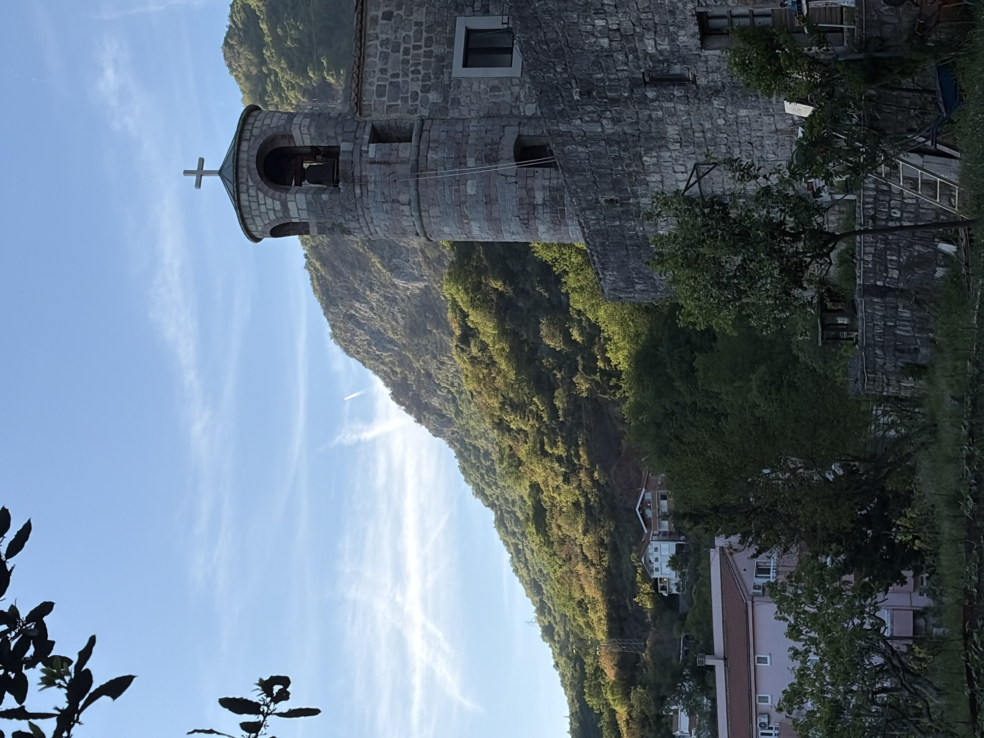 The image displays a scenic view, likely from Budva, Montenegro, featuring a stone building with a bell tower set against a backdrop of lush, green mountains and a clear blue sky. The building, constructed from grey stone blocks, has a distinctive bell tower topped with a Christian cross. Two arched openings for bells are visible in the tower, and windows of various shapes, including a square one with a dark frame, are present on the building's facade.
The mountains behind the structure are heavily forested with trees, and their peaks are bathed in sunlight, suggesting either morning or late afternoon. The sky above is largely clear, with a few wispy, white cirrus clouds stretching across it, and a faint contrail is visible higher up.
In the mid-ground, nestled into the mountainside, are several residential buildings with light-colored walls and characteristic orange or red tiled roofs. Trees and dense foliage are scattered throughout the scene, particularly in the foreground and around the stone building and residences, adding to the natural, verdant landscape. No specific activity or people are depicted in the static shot. There is no visible text in the image. The overall impression is one of a tranquil, picturesque setting in a mountainous coastal region.