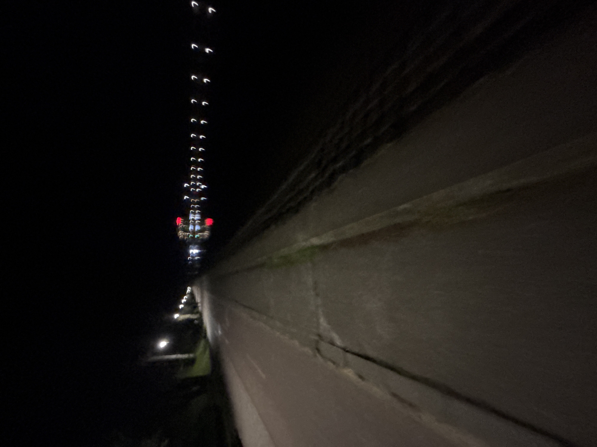 The image appears to be taken at night, possibly from a high vantage point, capturing a section of a concrete structure, likely part of a building or bridge, in the foreground. A line of blurry lights, ascending into the distance, suggests a street or thoroughfare running perpendicular to the concrete structure. The lights could be streetlights or vehicle lights. The environment is dark. While Plovdiv, Bulgaria, cannot be positively identified from this image alone, based on the architectural styles of buildings found in this city, the urban environment depicted in this photo could match that area.