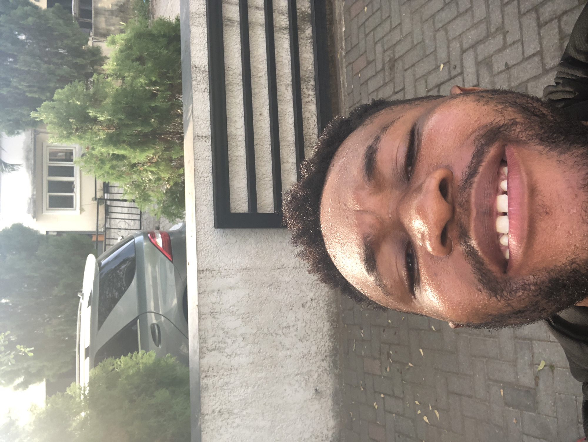 A close-up, angled view of a smiling man is featured in this photograph, taken outdoors. The man has a dark complexion, a short beard, and his eyes are partially closed. The skin on his face appears to have moisture.
In the background, there is a light gray wall with a black metal gate in front of it. To the left of the wall, greenery is visible, including a tree and the rear of a silver or gray SUV. The ground is paved with gray brick.