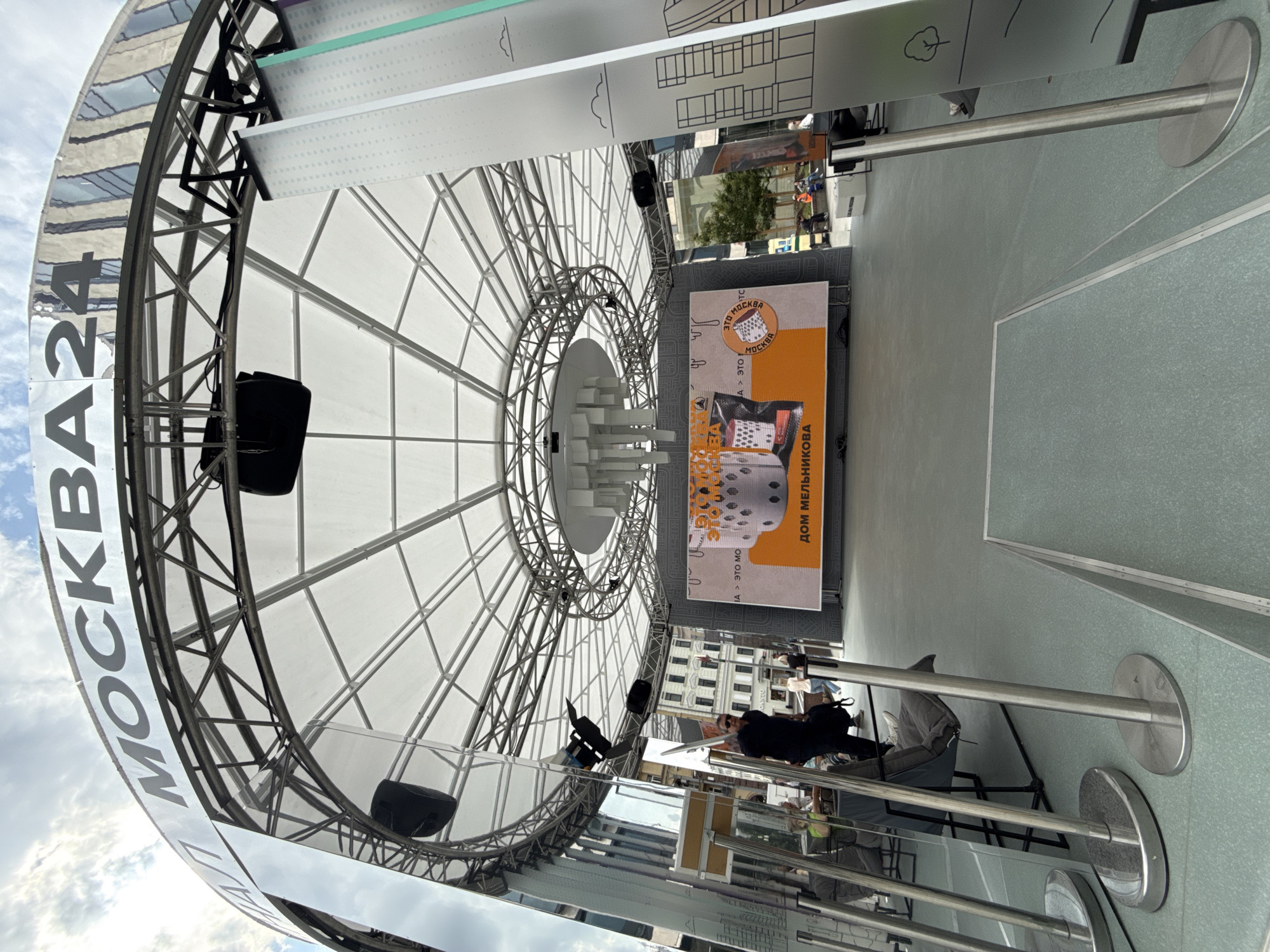 An outdoor stage area is set up under a white, tent-like roof supported by a metal truss structure. The words "МОСКВА24" are visible in large, black lettering on a silver, curved banner above the stage. A large LED screen displays an image featuring architectural elements and text, including "ДОМ МЕЛЬНИКОВА".
Several people are present in the scene: a man wearing sunglasses and a dark shirt stands near a reflective structure on the left. In the background, other individuals are seated or standing, some appearing to be workers in safety vests. A few empty chairs with cushions are positioned in front of the reflective elements. The ground is a light green surface, and metal stanchions with reflective poles are arranged in the foreground. The overall environment suggests an urban setting in Moscow, Russia, possibly a public square or event space.