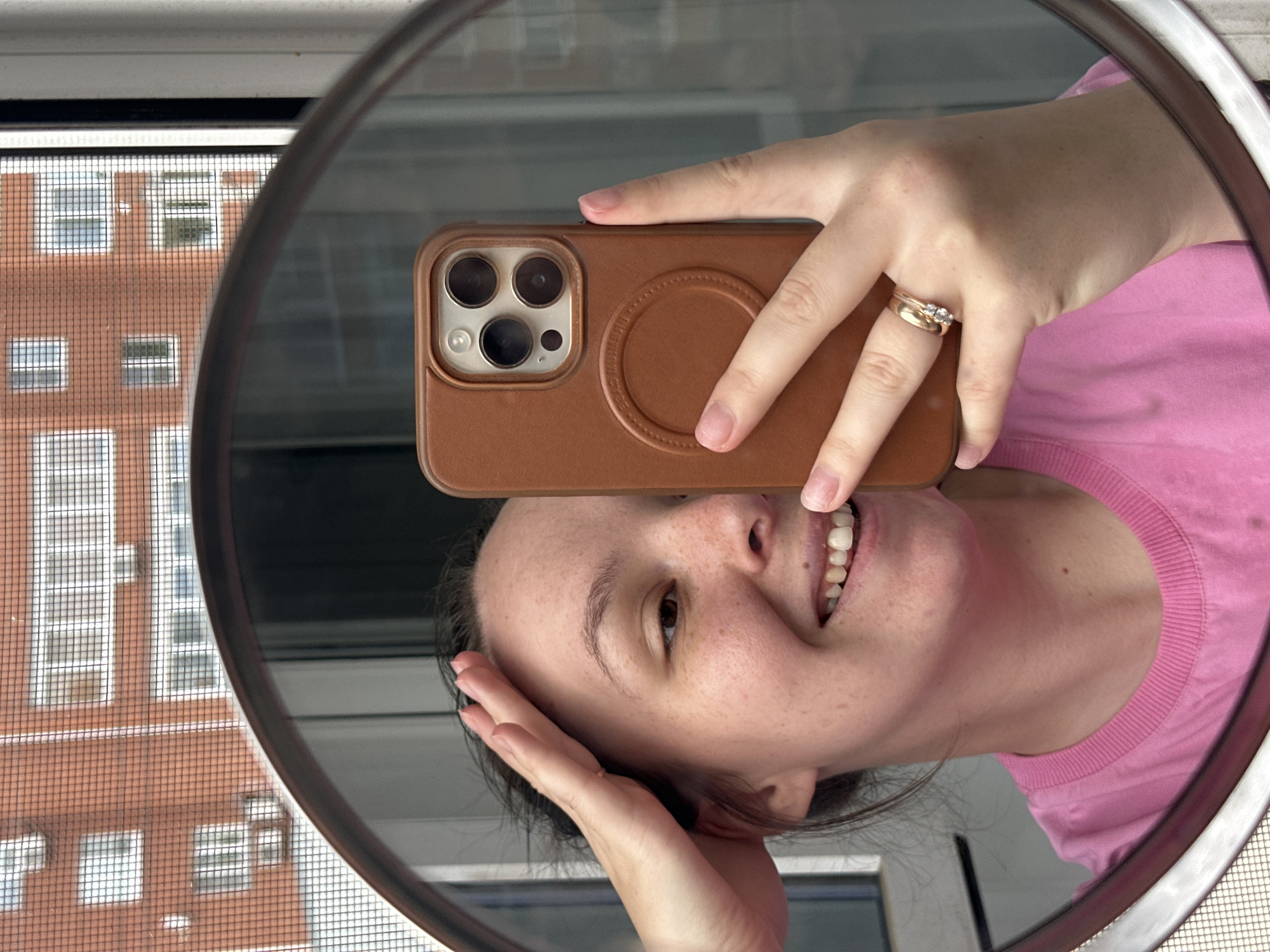 A young woman with dark hair and fair skin is taking a mirror selfie in a bathroom. She is wearing a pink t-shirt and gold rings on her left hand. Her right hand is holding a brown iPhone with a matching case. The mirror reflects the woman, her phone, and a window with a view of an apartment building. The apartment building is made of red brick and has white window frames. The window is covered by a screen. The woman is smiling and appears to be looking at her phone. The image is brightly lit, and the focus is on the woman and her phone. The location is the city of Chaikovsky, Russia.