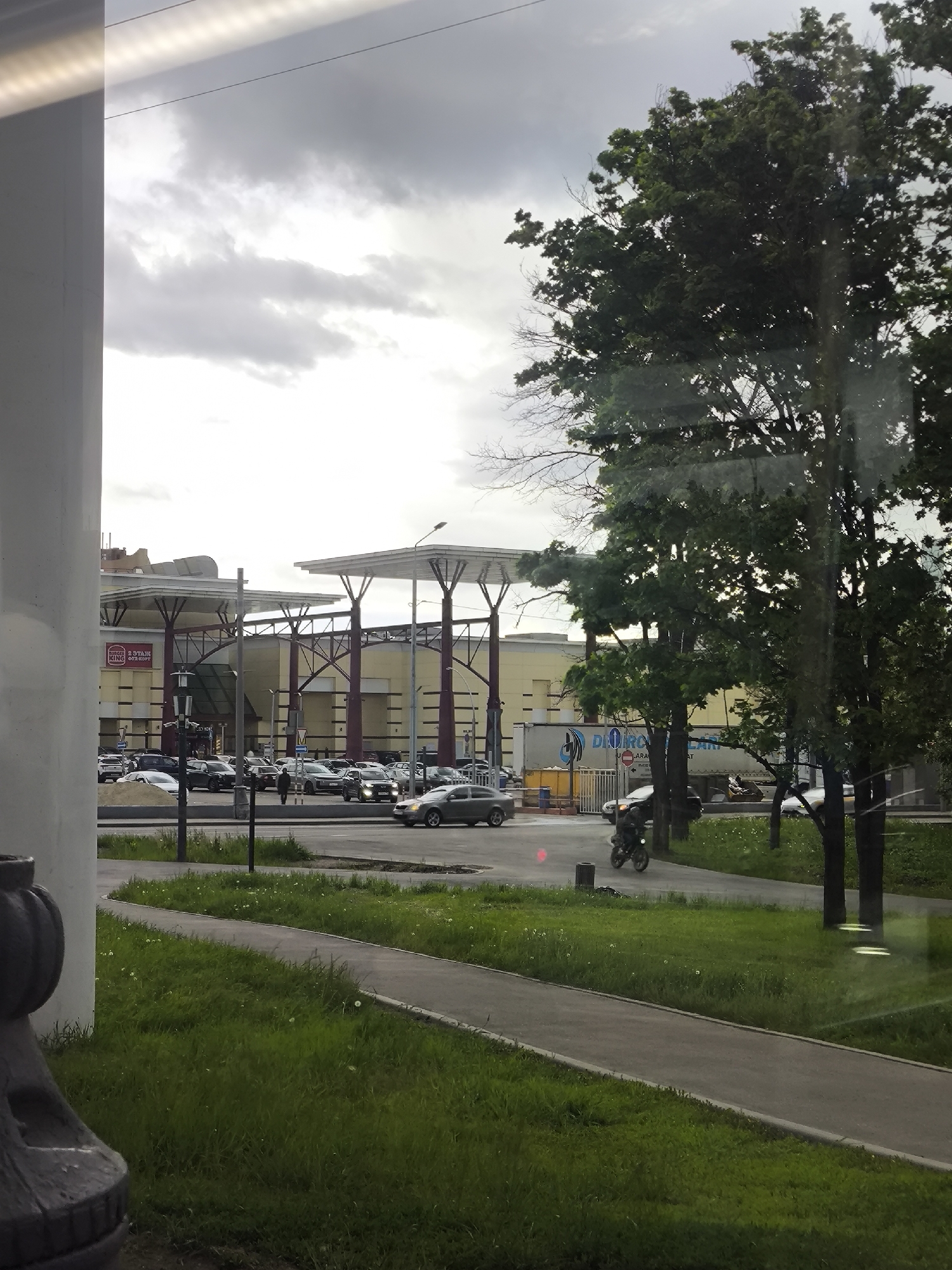 The image captures an outdoor scene in an urban environment. A gray paved walkway bordered by green grass runs along the foreground, transitioning into a street with active vehicle traffic. Behind the street is a large, tan-colored building with notable architectural features, including a high roof supported by multiple columns. The parking lot in front of the building is occupied by a number of vehicles. Amidst the traffic, a single motorcycle is visible. The sky is overcast with gray clouds. In the foreground and right side of the image, trees are present, partially obscuring the view. The perspective suggests the photo was taken from inside a building or vehicle, looking outwards.