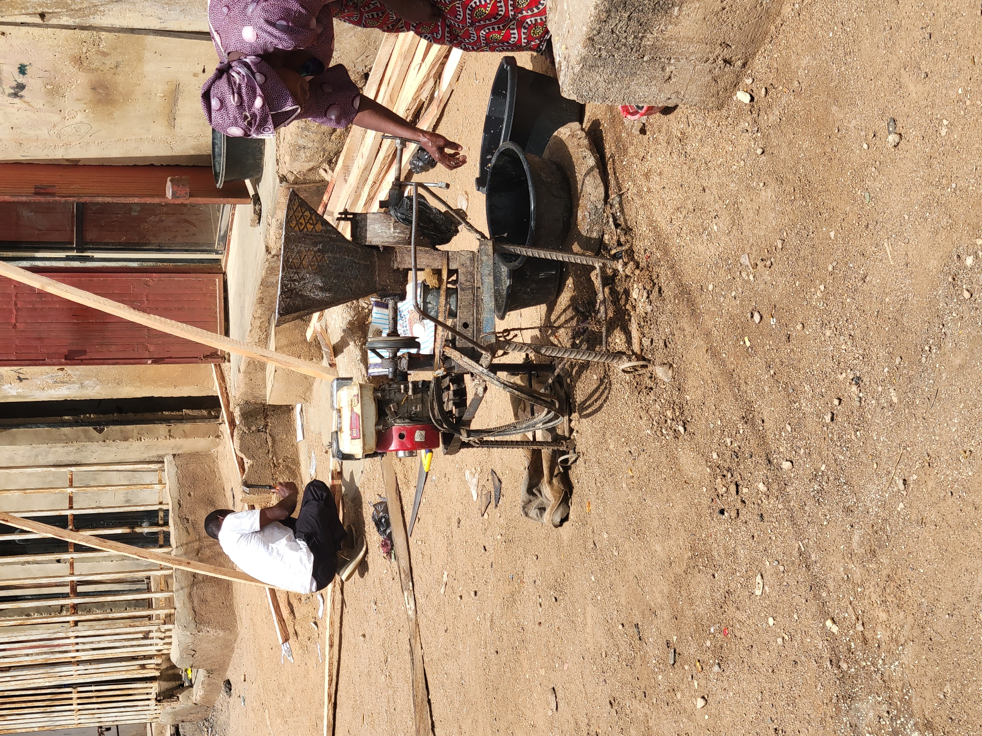The image depicts an outdoor scene, possibly a residential or commercial area, in Jos, Nigeria. The ground is predominantly sandy and earthy, with scattered small stones and debris.
Two individuals are visible. One is a woman wearing a patterned headwrap and dress, standing near a milling machine. Her arm is extended towards the machine, and she appears to be processing grains. The second person is a man, seated and working on a wooden structure. He wears a white shirt and dark trousers, and he is using a hammer.
The most prominent object is a milling machine. Two black buckets are placed next to it. The milling machine is powered by a small red gasoline engine.
In the background, there are buildings with painted walls and a metal gate. Wooden planks are scattered around the scene, suggesting construction or carpentry work.