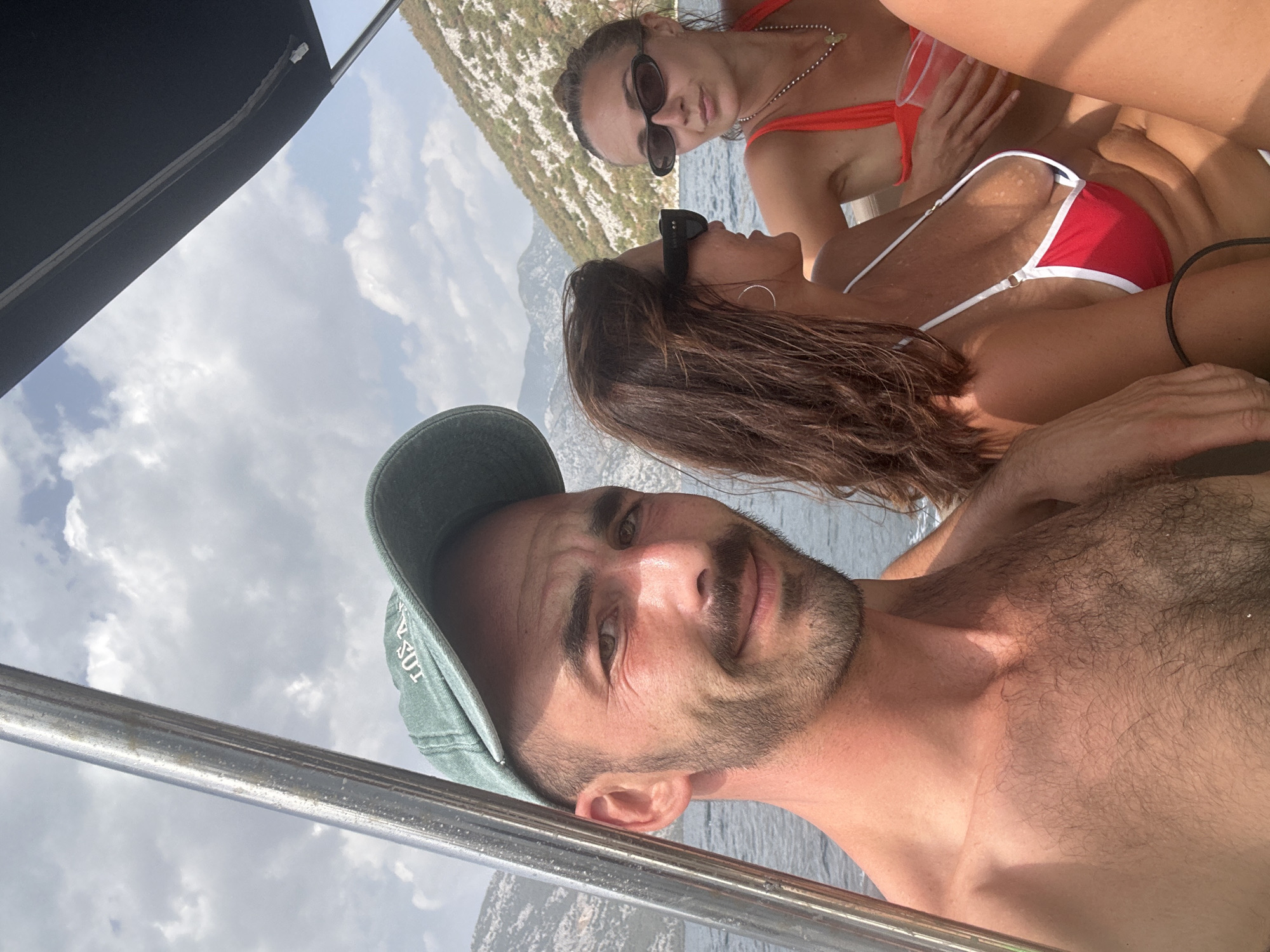 Three individuals, two women and one man, are seated on a boat in the waters of Perast, Montenegro. The man, in the foreground and to the left, wears a green baseball cap and has a mustache and beard. One woman, in the center, has long brown hair and wears a red and white bikini top. The second woman, on the right, has her hair pulled back and wears a red bikini top and sunglasses. They appear to be enjoying a sunny day on the water, with mountains visible in the background. The boat has a metal railing on the left and a dark canopy overhead.