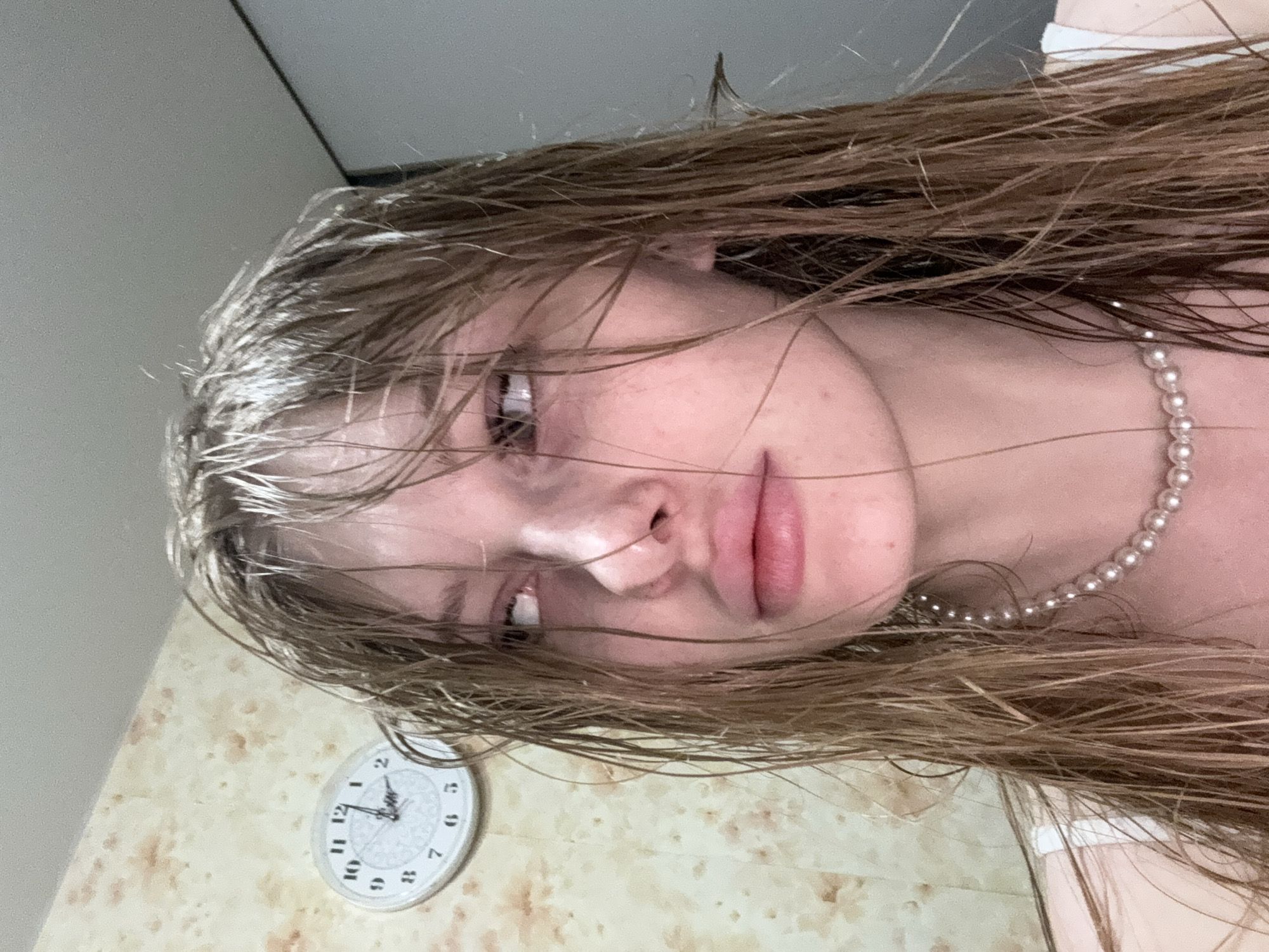 A close-up, head-and-shoulders photograph depicts an individual with long, wet, light brown hair hanging around their face. The individual's eyes are rolled upwards and to the right, exposing significant sclera, and their lips are slightly parted. They wear a single strand of small pearls around their neck, with a white strap of clothing visible on their left shoulder. The background on the left side features a patterned wall with light beige and irregular brown/orange markings. Mounted on this wall is a round, white analog clock with black Arabic numerals, displaying approximately 12:12. The background on the right consists of a plain, light grey wall or surface, marked by a sharp, dark vertical line indicating a corner or frame. The scene is located in the city of Perm, Russia.