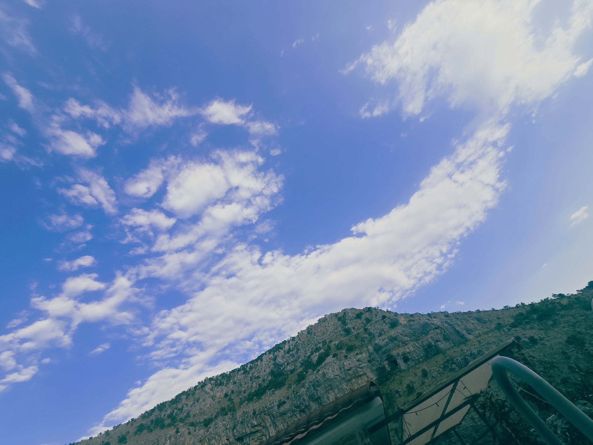 The image shows a rocky cliff face on the left, extending upwards from the bottom left corner and diagonally across the frame. The cliff is covered in sparse vegetation. In the foreground, a portion of a metal railing and a canopy structure are visible, suggesting a viewpoint or seating area. The right side of the image is dominated by a bright blue sky with scattered white clouds, some of which are wispy and elongated. The overall impression is that of a scenic overlook, likely in a mountainous or hilly area. This scene is consistent with various locations in Podgorica, Montenegro, known for its proximity to natural landscapes with cliffs and mountains.