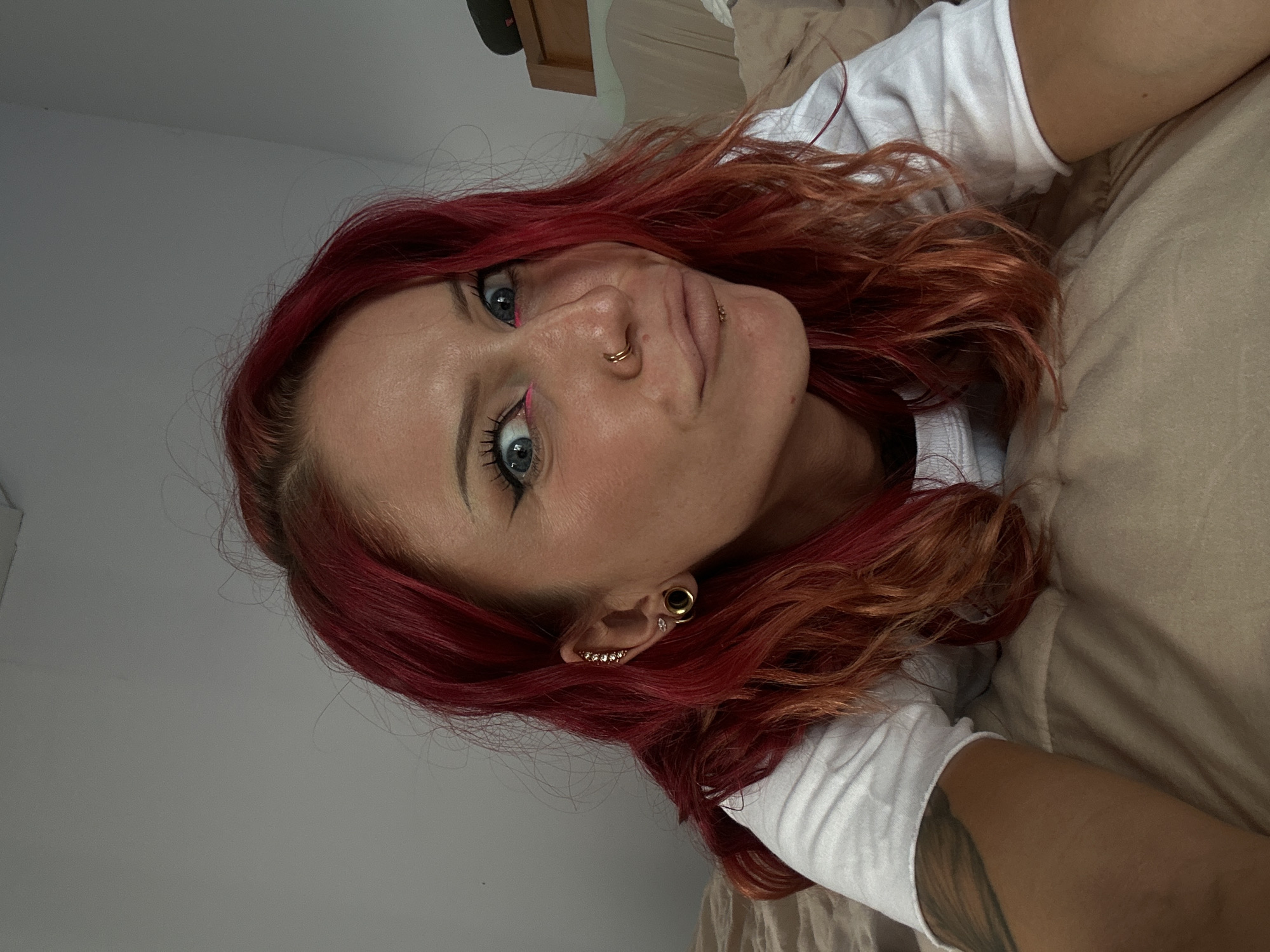 A woman with red and auburn dyed hair is lying on a beige bed, angled towards the viewer. She has light blue eyes with pink eyeliner and multiple piercings, including a double nose ring, lip piercing, and a stretched earlobe with various earrings. She's wearing a white t-shirt. The background consists of a light gray wall and a partially visible wooden cabinet. The image appears to be taken indoors, potentially in a bedroom. There is no clear evidence to confirm the location is specifically North Kuta, Indonesia.