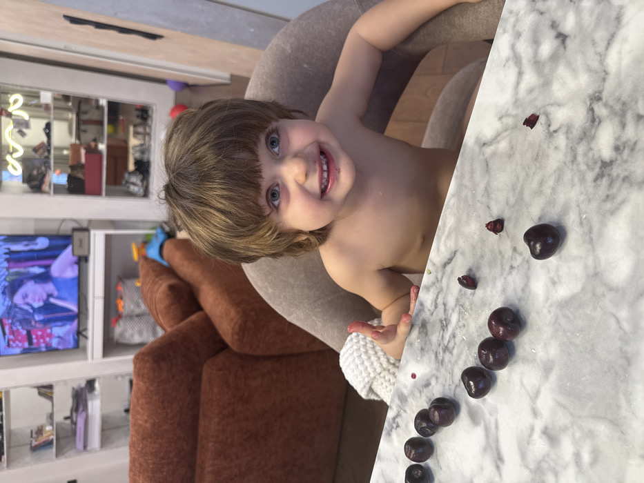 Location: Indoor domestic setting, in the city of Unknown, Unknown.
A light-skinned toddler, with brown, bowl-cut hair and blue eyes, is seated shirtless in a light beige upholstered chair at a white marble-patterned table. The toddler's mouth area and fingers of the left hand are stained dark red, consistent with cherry juice. The toddler is looking directly forward and smiling broadly, with teeth visible. Scattered on the table in front of the toddler are approximately eight whole dark red to purplish cherries and several fragments of cherry pits and flesh. A white knitted textile is draped over the left armrest of the toddler's chair. In the background, a large, rust-colored upholstered sofa is partially visible. Behind the sofa, a white wall unit features a television displaying an image of a woman, flanked by white shelving units. The left shelving unit contains various items, while the right unit, enclosed by glass doors, displays shoes and other objects. A Marshall brand speaker is situated below the television.