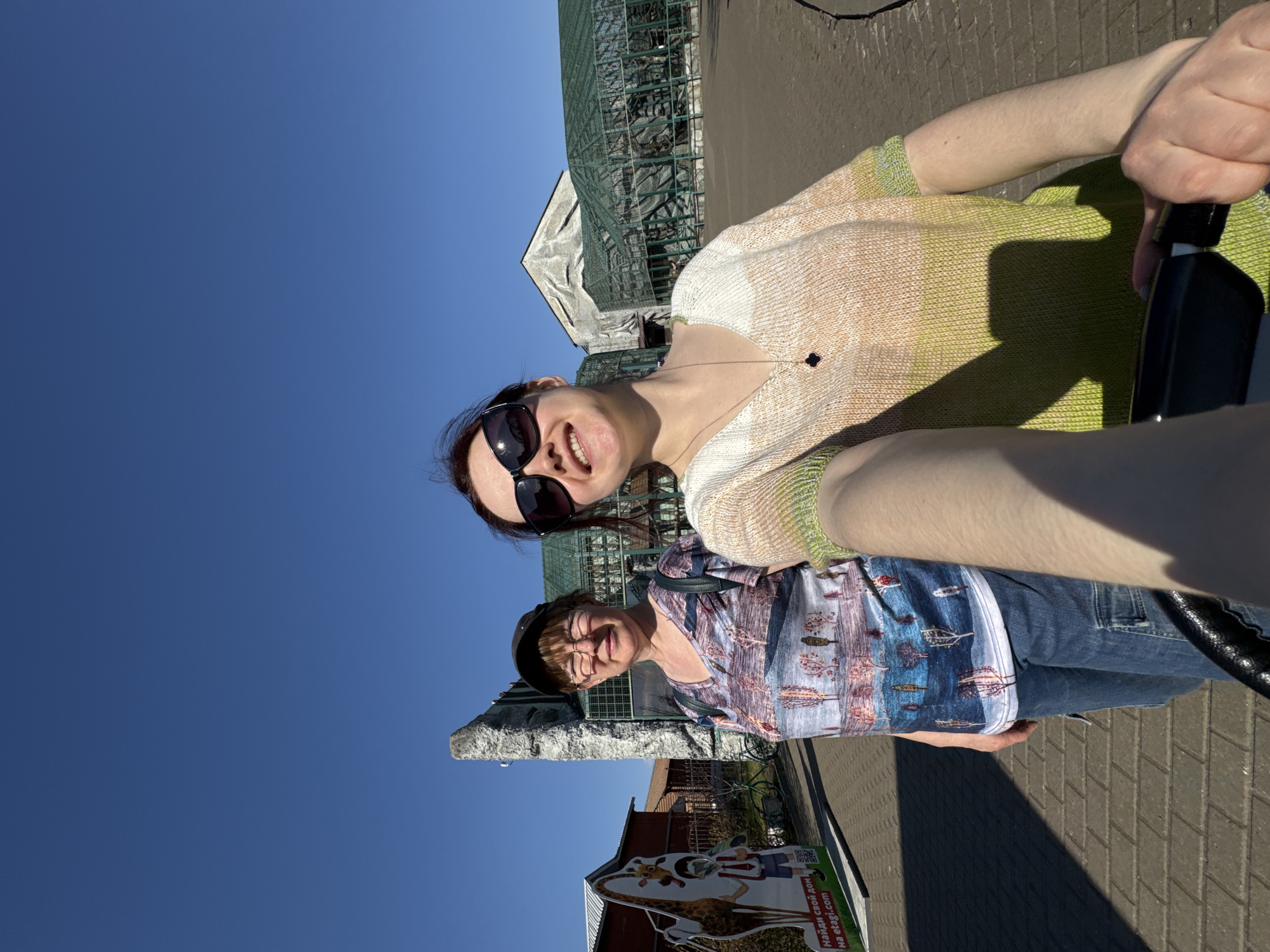 The image shows two women taking a selfie outdoors. The woman in the foreground is wearing sunglasses and a light-colored sweater. Behind her is an older woman with glasses and a patterned shirt. They are standing in front of a green metal structure, possibly a greenhouse or aviary. In the background, there is a statue and signage. The location appears to be the Izhevsk Zoo in Russia.