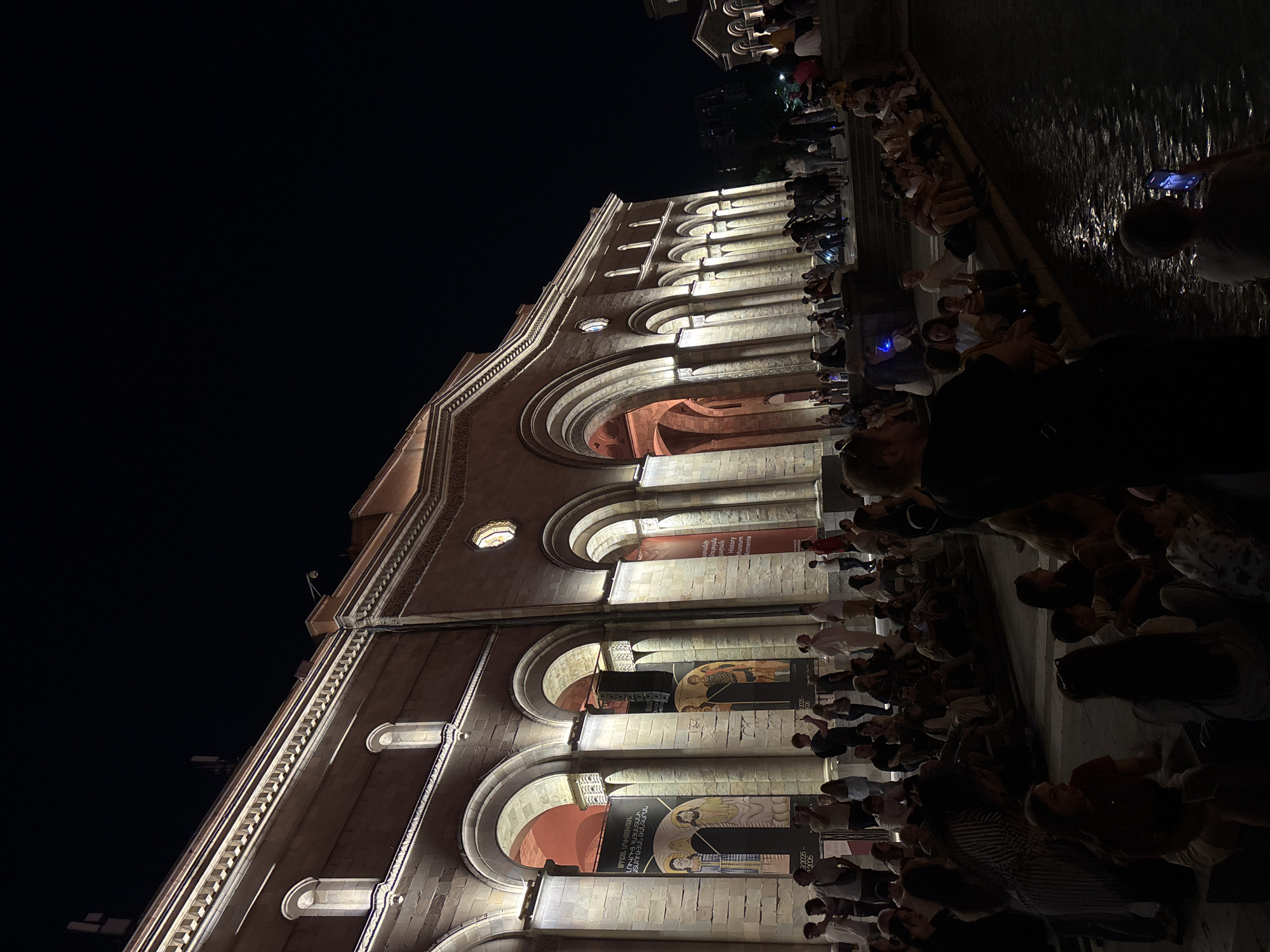 The image captures a nighttime scene in Yerevan, Armenia, outside the History Museum of Armenia. A large crowd of people is gathered, many sitting on steps and the edge of a water feature. The museum building, illuminated by warm lights, dominates the background with its classical architecture, featuring arched entrances and decorative stonework. Banners are hung within the arches, displaying images and text in Armenian. The overall atmosphere suggests an evening event or gathering.