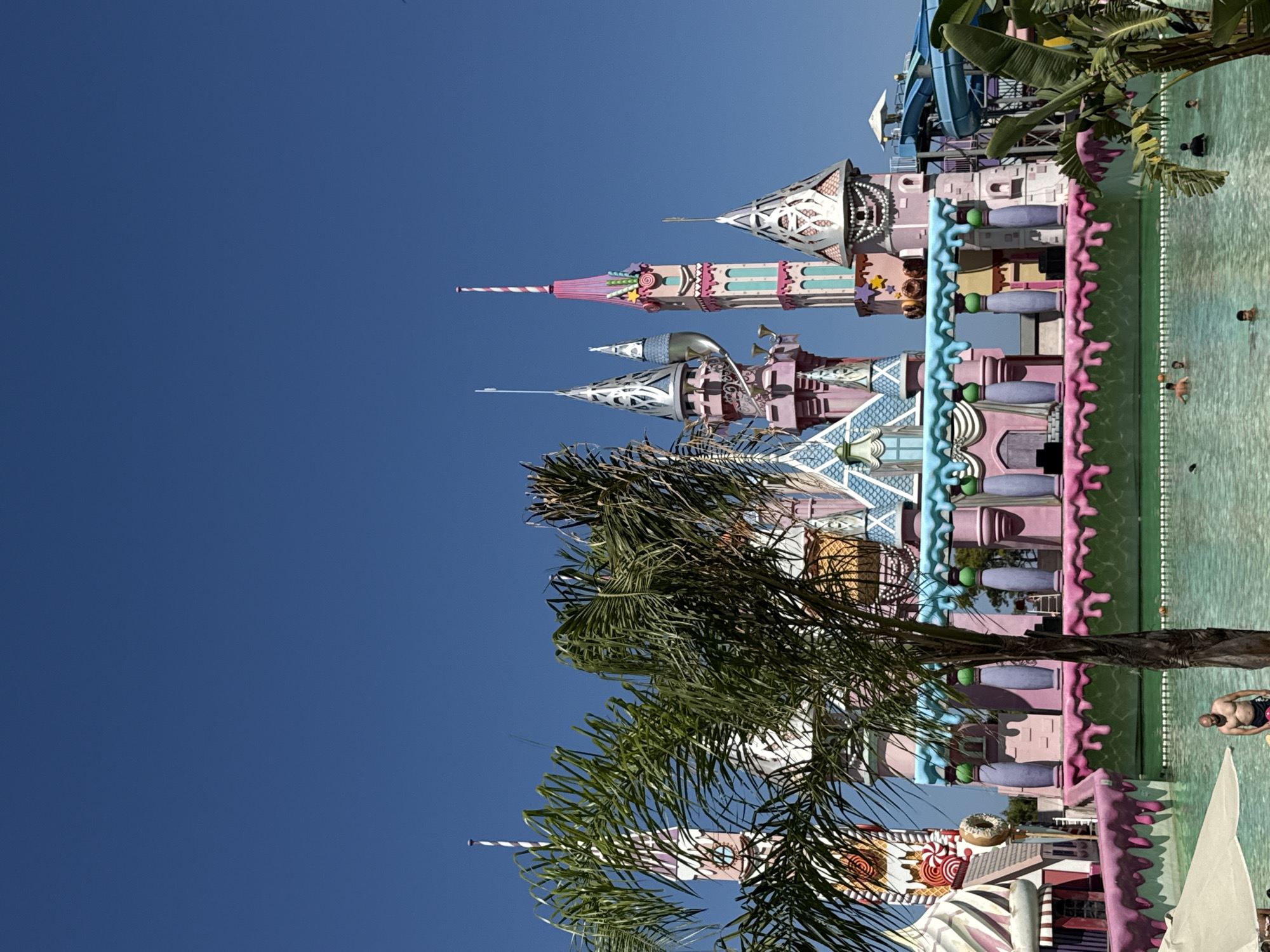 A low-angle, eye-level, outdoor, full shot captures a candy-themed amusement park, likely located in Bodrum, Turkey. In the foreground, a man is standing in a shallow, clear turquoise pool, his chest and head visible above the water. He is partially submerged, facing the viewer with a neutral expression. The pool extends towards the background, where a large, whimsical castle-like structure dominates the scene. The castle is painted in vibrant pinks, purples, and blues, with elaborate turrets, spires adorned with candy motifs like lollipops and donuts, and a dripping frosting-like trim. Palm trees with fronds obscuring parts of the castle are present on the left and right sides of the frame. The sky above is a clear, deep blue. Several other individuals are visible in the pool, appearing to swim or lounge in the water. A blue waterslide structure is visible to the right of the castle. The overall impression is that of a brightly colored, playful environment, likely a water park or theme park.