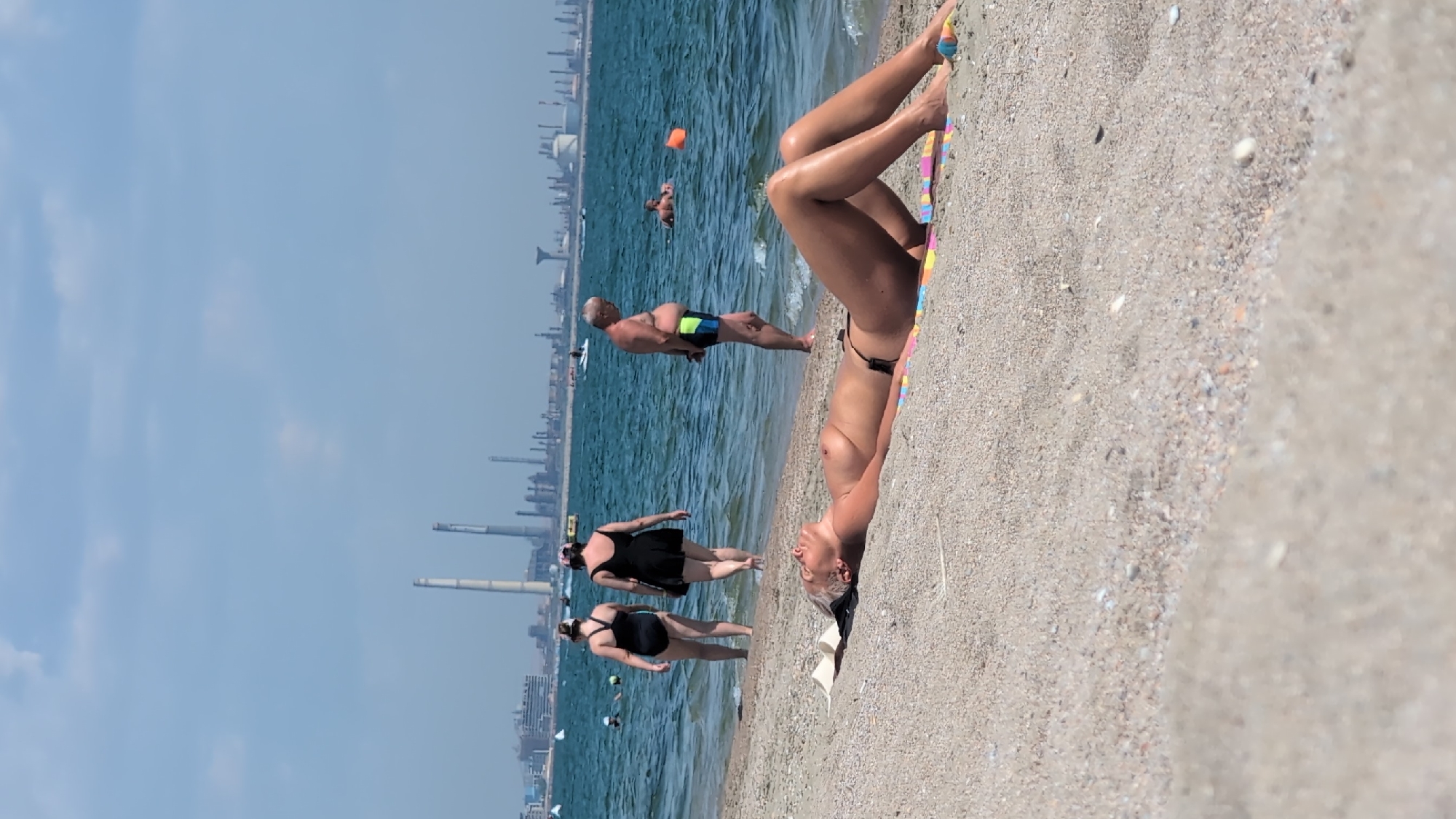 This image depicts a beach scene in Constanța, Romania, likely in the Mamaia area due to the industrial skyline in the background. In the foreground, a woman is sunbathing on the sand, lying on her back with her eyes closed and her body exposed. She is wearing a black bikini bottom and a colorful striped towel beneath her. In the middle ground, the Black Sea stretches out, with several people visible. Two women in black swimsuits are walking towards the water, one with her back to the camera and the other facing away. A man in swim trunks is standing by the water's edge, and another man is swimming further out in the sea. An orange buoy is visible in the water. In the background, a sprawling industrial complex with numerous smokestacks and buildings lines the horizon, indicative of Constanța's industrial port. The sky is clear and blue with scattered clouds.