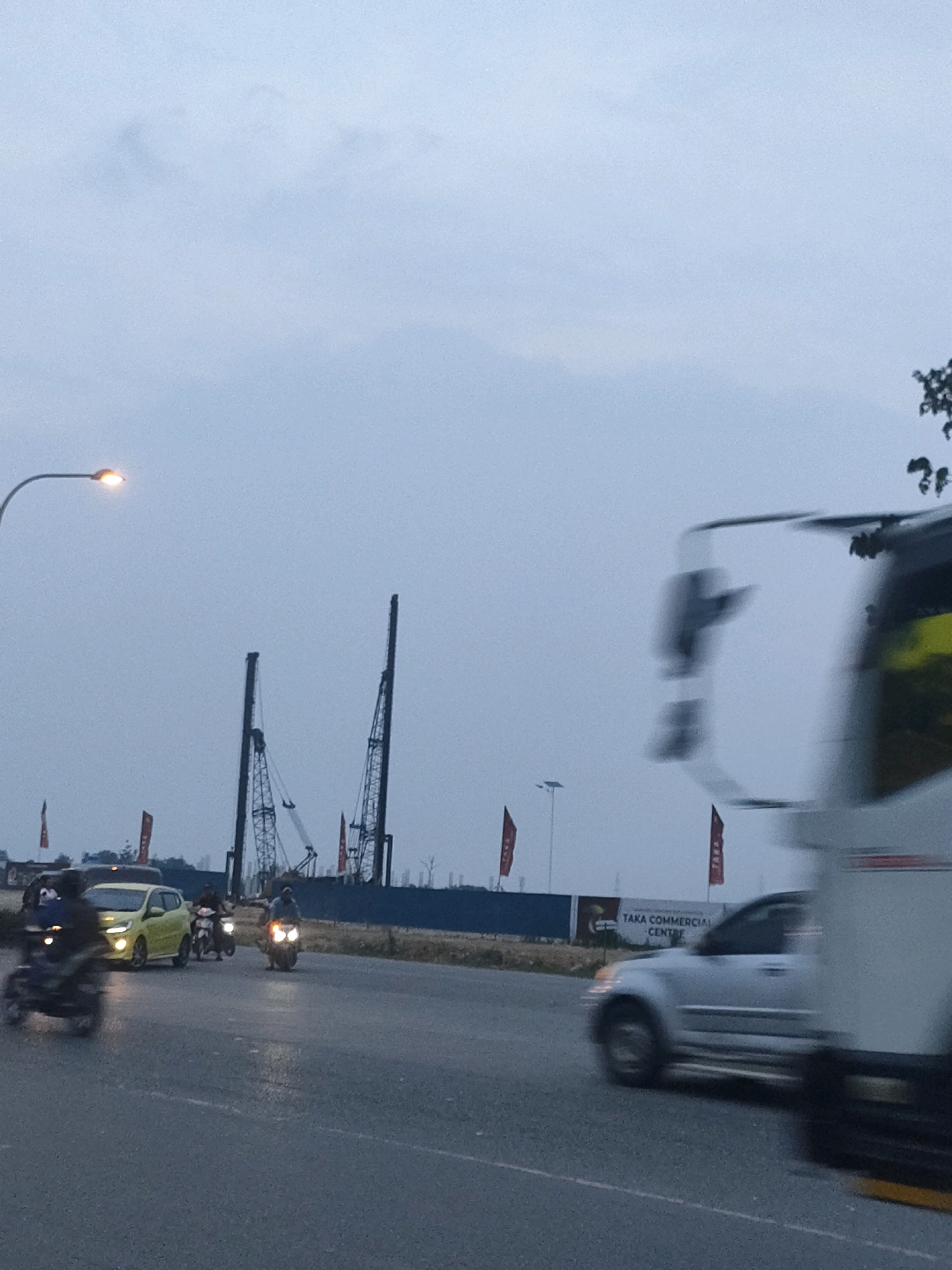 The image depicts a street scene in Batam, Indonesia, likely during the late afternoon or early evening, given the low light conditions and illuminated streetlights. In the foreground, a blurry white vehicle, possibly a bus or large truck, dominates the right side of the frame, its motion suggested by the streaking effect.
On the road, several motorcycles are visible. One motorcycle, ridden by a person in dark clothing, is in motion on the left side of the road. Further down the road, a yellow car is stopped or moving slowly, with two motorcycles nearby. One motorcycle is to the left of the car, and another is in front of the car, with its rider wearing a helmet.
In the background, there is a construction site with two large cranes. A fence or barrier encloses the site, and a billboard with the text "TAKA COMMERCIAL CENTRE" is visible. Red flags are planted along the fence line. The sky is overcast and appears a muted blue-grey. A street lamp with an orange glow is positioned on the left side of the image, illuminating a section of the road. Trees are sparsely visible on the horizon.