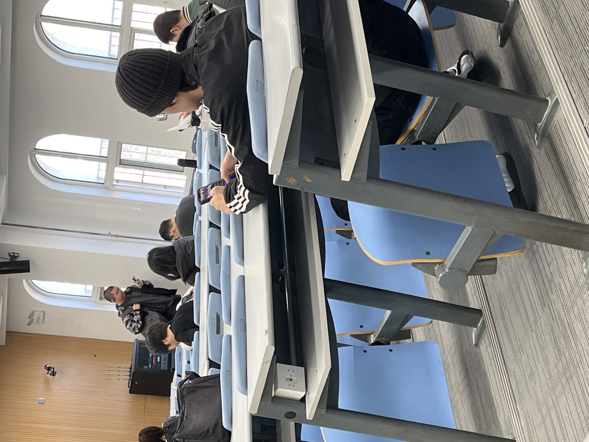 The image depicts a classroom setting. A row of students is seated at desks with light blue seats and white writing surfaces. At least seven individuals are visible, most focused on electronic devices. Some are looking at cell phones, while others appear to be reading documents.
The classroom is well-lit with natural light coming through arched windows. The interior includes light-colored walls and a grey striped floor. An electrical outlet is visible on one of the desks. In the background, there is a wooden-paneled wall and what appears to be sound or electronic equipment.
I cannot verify whether this is in Changchun City, China. I do not have access to that information.