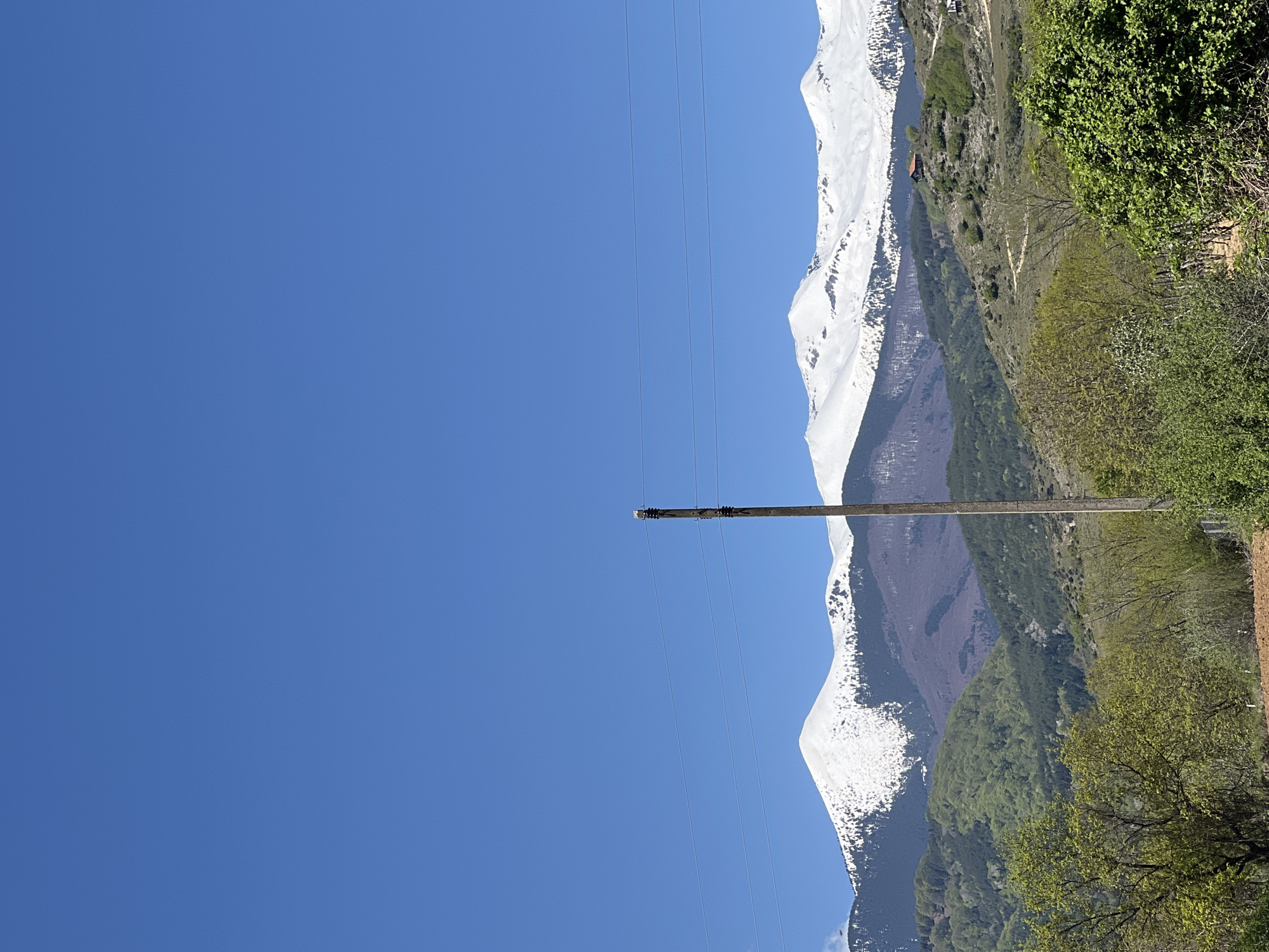 The image depicts a vertical view of a landscape in Kornitsa, Bulgaria, featuring a clear, bright blue sky dominating the left side. A tall wooden utility pole stands in the center, with electrical wires extending horizontally across the sky. To the right, snow-capped mountains form the backdrop. The lower slopes of the mountains are covered with green vegetation and trees. A brown dirt road is visible along the lower right side of the image, adjacent to the greenery. A small red-roofed building is discernible on the mountainside. There is no visible presence of people, subjects, actions or interactions.