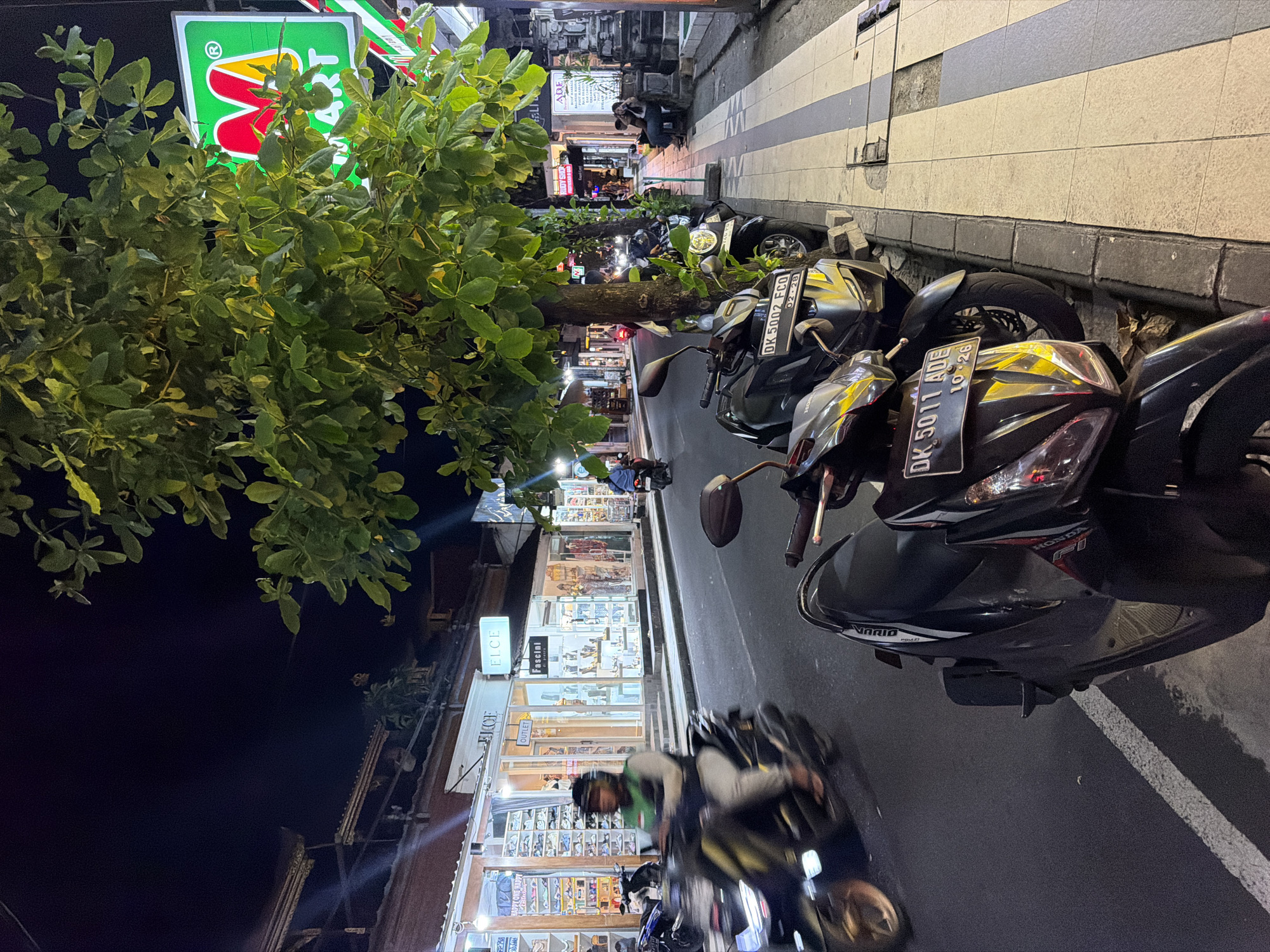 This vertical image depicts a lively street scene at night in Kuta, Indonesia. The foreground is dominated by the dense foliage of trees and several parked motorbikes along a paved sidewalk. The street and shops are brightly illuminated by artificial lighting, contrasting with the dark night sky.
A large, partially obscured sign for what appears to be a convenience store, featuring a green background and a red 'M' shape, with "ART" visible, stands out behind the trees. Below it, a row of commercial establishments lines the street, their storefronts glowing with displays of various goods. Visible text on these shops includes "ELCE," "OUTLET," "Fascini," "RUDY SHO," and "HAPPY ONE HAPPY."
Several motorbikes, including models like a Honda Vario, are parked on the sidewalk. Their license plates, "DK 5002 FCQ" and "DK 5011 ADE," indicate the location as Bali. A person on a scooter, blurred by motion, is visible in the foreground, suggesting ongoing traffic. Other individuals are seen walking or crouching along the tiled sidewalk further down the street. The scene conveys a bustling commercial atmosphere typical of an evening in a popular area.
