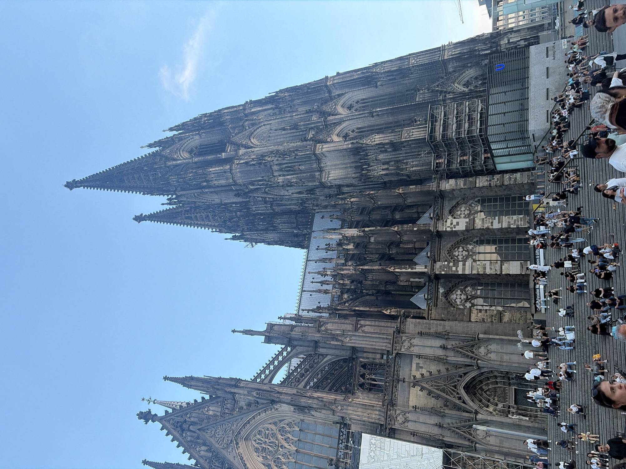 The image depicts a low-angle shot of the Cologne Cathedral in Cologne, Germany, against a clear blue sky. The imposing Gothic structure dominates the frame, with its intricate stonework, pointed spires, and large stained-glass windows visible. Scaffolding is present on a section of the cathedral, suggesting ongoing maintenance or construction.
At the base of the cathedral, a wide set of stone steps is populated by a crowd of people. They are casually dressed and appear to be relaxing, socializing, or observing the cathedral. Some are sitting on the steps, while others are standing or walking. The diverse group includes individuals of various ages and ethnicities.
The environment is an urban public space, likely a plaza or square in front of the cathedral. The lighting indicates it is daytime, with soft sunlight casting shadows. The overall atmosphere appears to be one of tourism and leisure, with many visitors taking in the famous landmark.