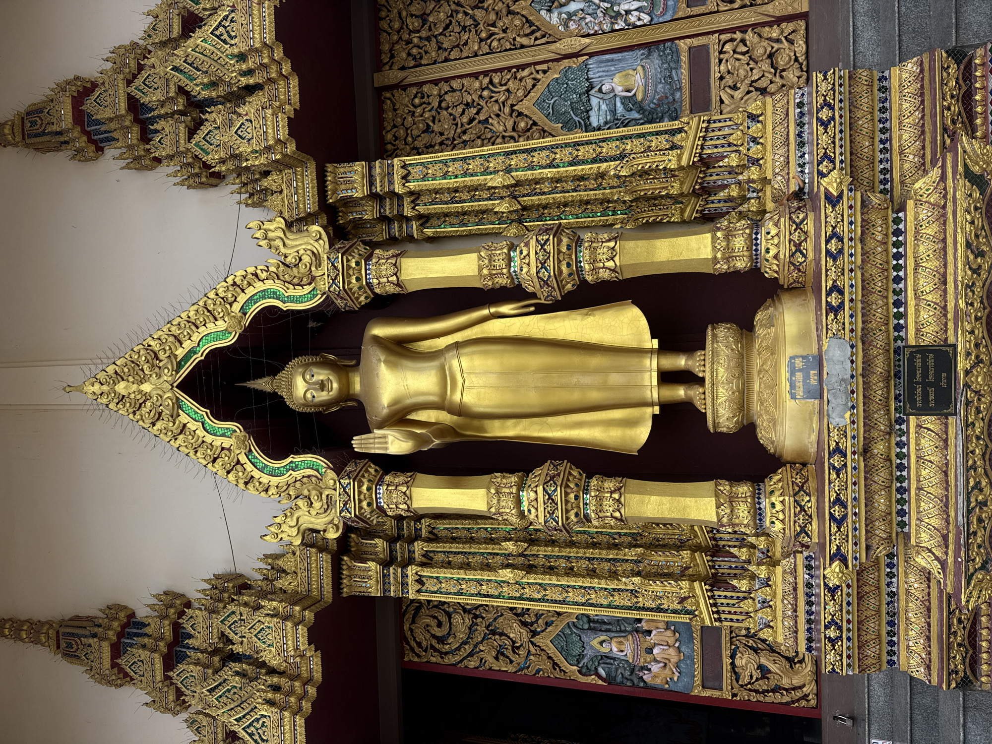 A golden statue of the Buddha stands prominently in the center of the image, positioned within an ornately decorated alcove. The Buddha is depicted in a standing posture, with its right hand raised in a gesture of blessing or protection. The statue is adorned with a flowing robe and a halo-like headdress. The alcove itself is a masterpiece of craftsmanship, featuring intricate golden carvings, colorful mosaic inlays, and elaborate architectural elements that frame the Buddha. Flanking the central statue are richly decorated pillars and walls adorned with detailed bas-reliefs depicting scenes that appear to be of religious or mythological significance. The overall scene suggests a sacred space, likely a temple or shrine, characterized by opulence and devotion. The location is identified as Koh Samui District, Thailand.