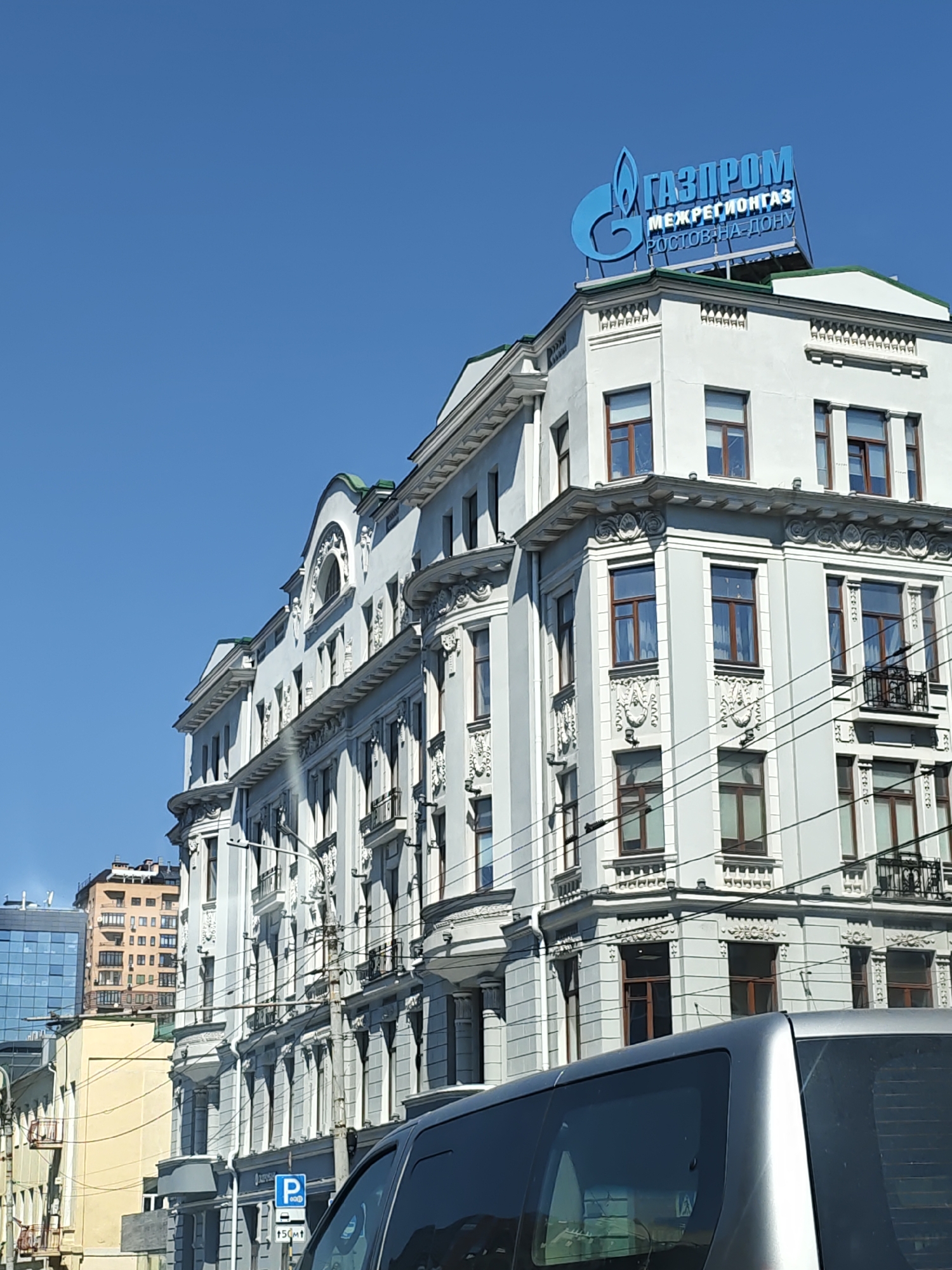 The image shows a building with the blue Gazprom logo and Cyrillic text on its roof. The text reads "ГАЗПРОМ" (Gazprom) on the top line and "МЕЖРЕГИОНГАЗ" (Mezhregiongaz) and "РОСТОВ-НА-ДОНУ" (Rostov-on-Don) on the lines below, identifying it as the Gazprom Mezhregiongaz Rostov-on-Don office. The building is white with ornate architectural details, including columns, cornices, and decorative moldings around the windows. It has multiple stories and numerous windows, many of which are dark-colored. Several electrical wires are visible in front of the building. The sky is clear and blue. In the foreground, the roof and side window of a silver vehicle are visible. In the background, other buildings of varying heights and architectural styles can be seen. The overall scene depicts an urban environment under a clear sky.