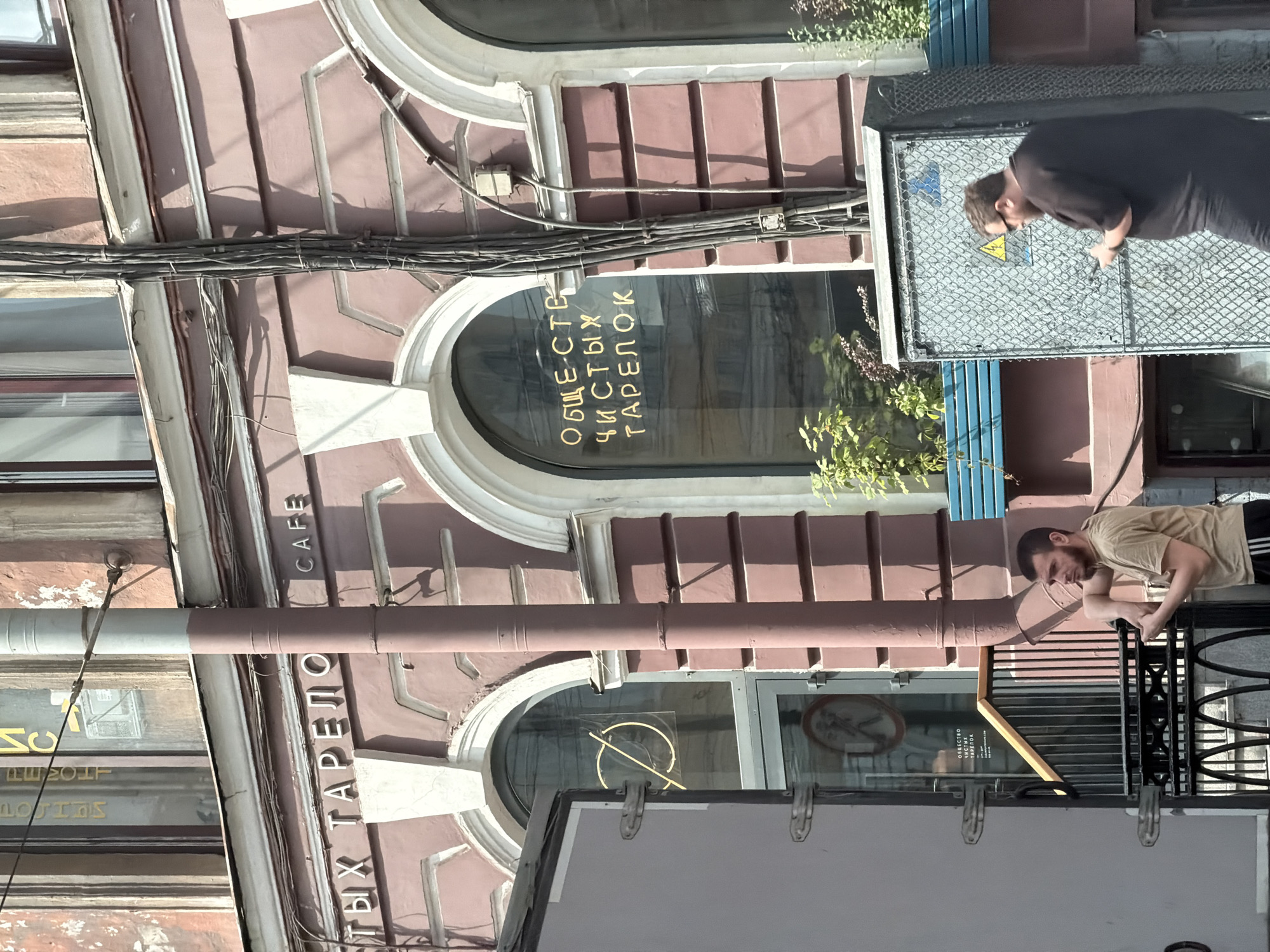 In Saint Petersburg, Russia, on a sunny day, an exterior shot focuses on a building with a pinkish-brown facade. Above an arched window on the right, the Cyrillic word "CAFE" is visible in white lettering. To the left of this, another arched window displays gold lettering that reads "ОБЩЕСТВО ЧИСТЫХ ТАРЕЛОК" (Society of Clean Plates). Further left, on the facade, the Cyrillic word "ТАРЕЛО" is partially visible. A dark grey pipe runs vertically up the center of the image. In the foreground, to the right, a man wearing a black t-shirt and sunglasses stands in front of a gray, chain-link-covered electrical box, looking down at his phone. To the left, another man in a tan t-shirt and black shorts stands near a black metal railing, his left arm resting on the railing. Sunlight casts shadows on the building.
