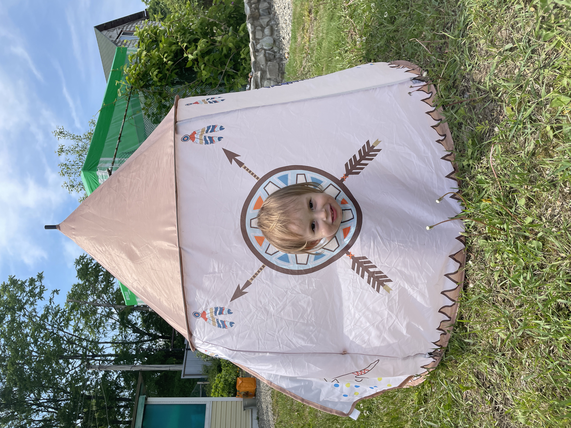 A color image shows a young child with blonde hair looking through a circular opening in a teepee-style tent. The tent is positioned outdoors on a lawn with green grass. The tent has a beige and white color scheme with a brown border and decorative patterns resembling arrows and feathers. In the background, there are trees, parts of buildings, and a patch of blue sky. This image was taken outside of Yasnoe, Russia.