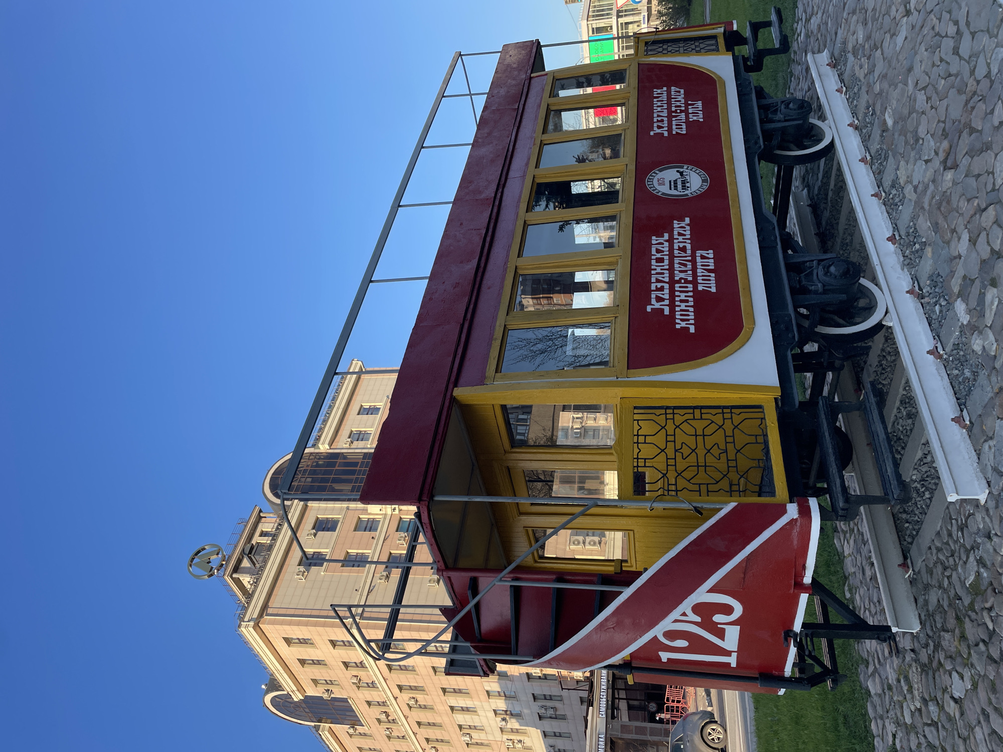 A color photograph depicts a vintage tram car in Kazan, Russia, under a clear blue sky. The tram is painted primarily in red, yellow, and white. It has a series of rectangular windows with yellow frames. Russian text is painted on the red portion of the tram. The number "125" is prominently displayed in white on the front of the tram. The tram sits on a short section of railway track, which is laid on a cobblestone surface. Behind the tram, a large building with multiple floors and many windows is visible. At the top of the building, a circular symbol is mounted. A modern car is also visible in the background.