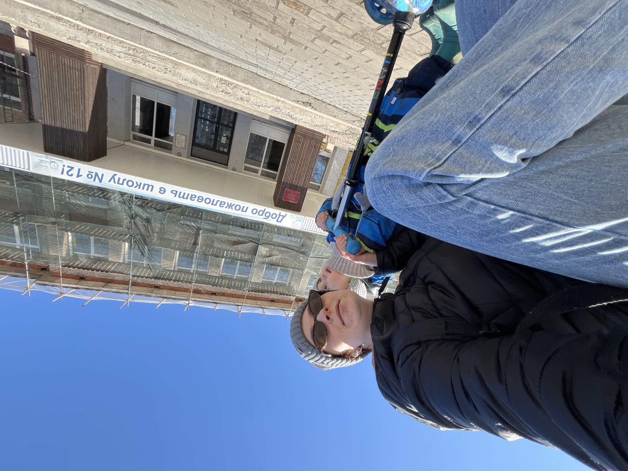 A low-angle shot captures a person wearing a black jacket and grey beanie, sunglasses, and blue jeans, seemingly holding a child in front of them. The child is wearing a blue and black striped jacket and is holding a scooter. A building under renovation is visible in the background, with scaffolding and netting covering part of the facade. A sign with Cyrillic text is attached to the building; it says "Welcome to school No. 12!". The location is Chaikovsky, Russia. The sky is clear and blue.