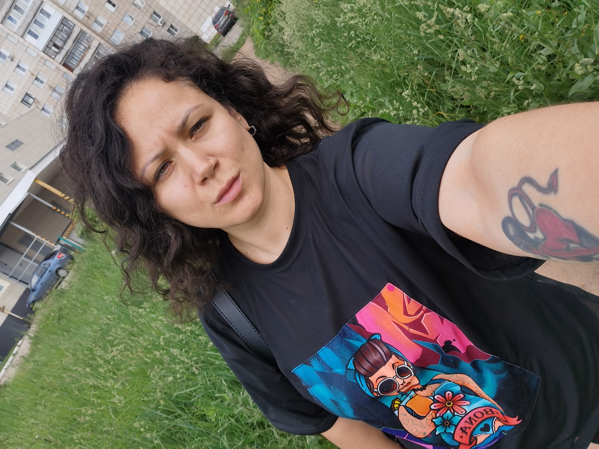 A woman with dark curly hair takes a selfie outdoors in front of a residential building and a grassy area. She is wearing a black t-shirt with a colorful graphic print of a person with sunglasses. A tattoo is visible on her arm. The background includes a multi-story apartment building, a road with a parked car, and a patch of overgrown grass with small white flowers. The scene suggests a casual outdoor setting in a residential urban environment.