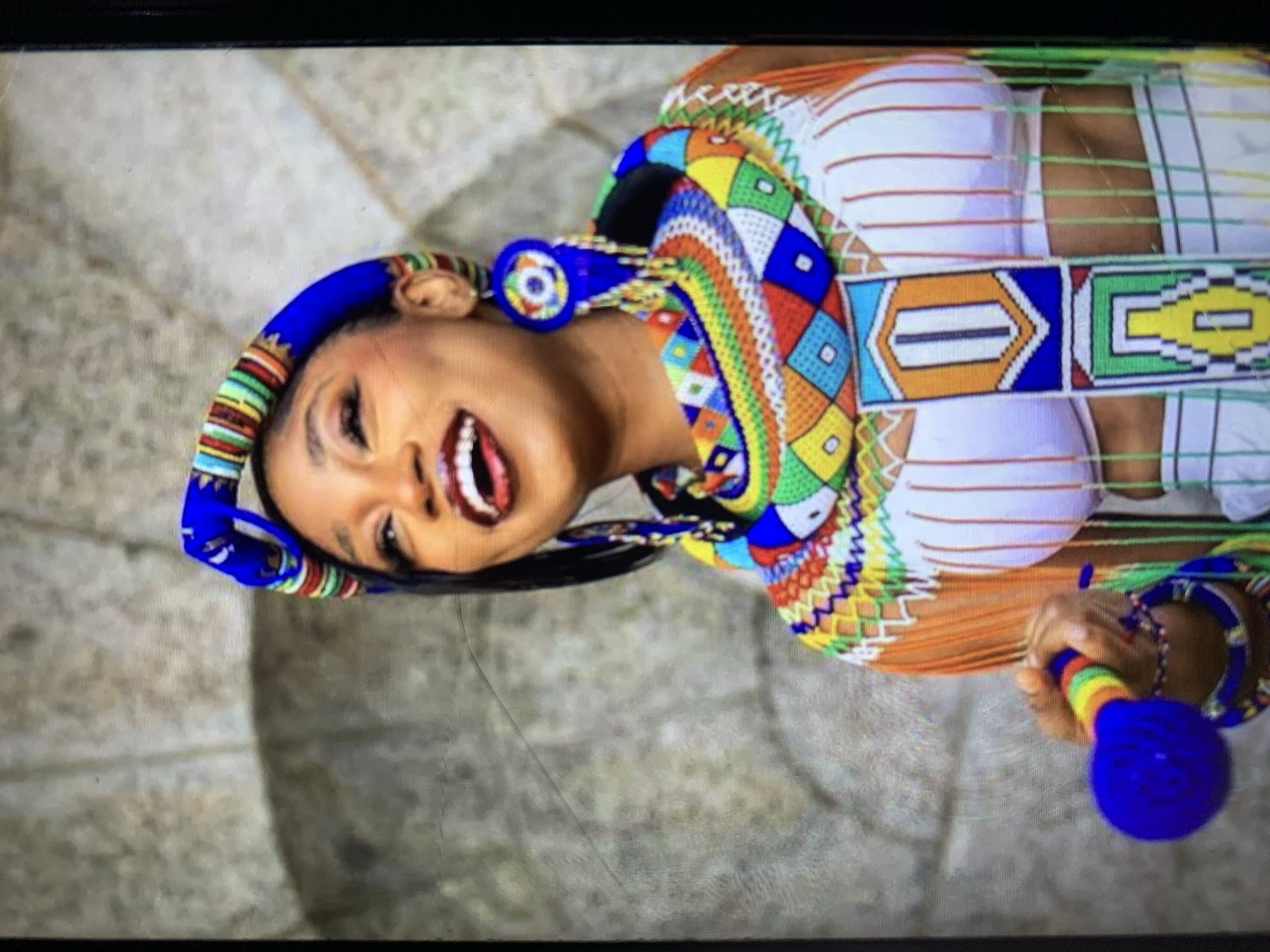 A smiling woman with a blue beaded headband and matching beaded earrings, necklace, and bodice stands in front of a circular, stone structure. She holds a blue beaded rattle in her right hand. The necklace and bodice are patterned with colorful geometric designs. Her white top has thin horizontal stripes. The location appears to be outdoors and the subject may be in New Stadium, Nigeria.