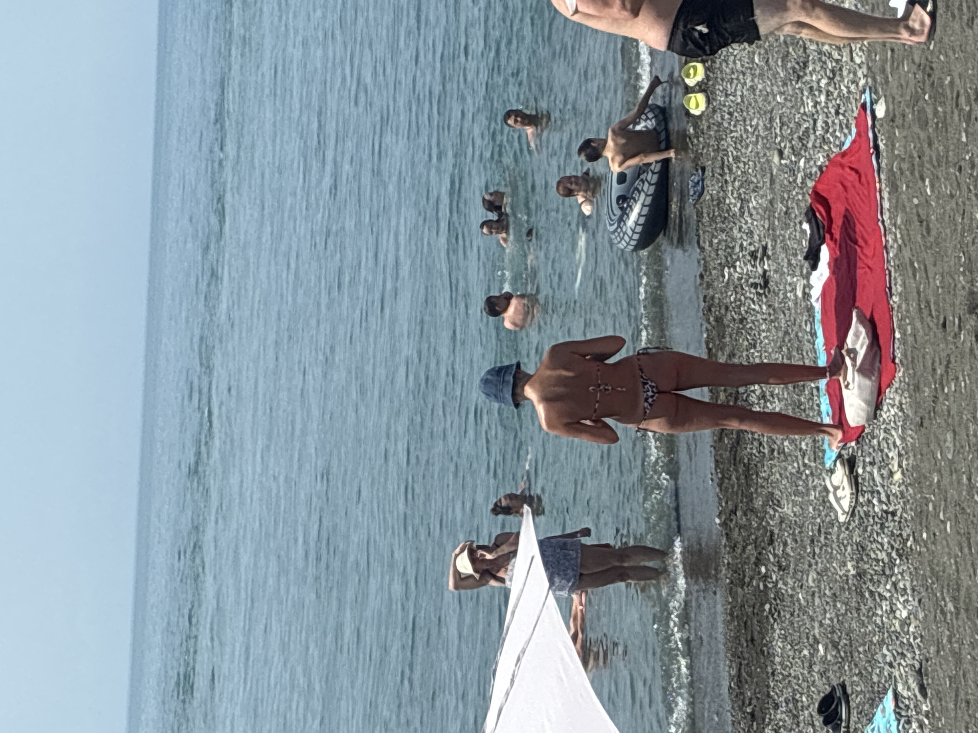 This is a medium shot of a beach scene in Sirius, Russia. The image is oriented vertically, showing the ocean on the left and the pebble beach on the right. Several people are in the shallow water, some swimming and others standing. In the foreground on the right, a woman in a bikini stands on the beach, facing away from the camera. A red and white towel is spread on the pebbles behind her, along with some flip-flops. Further down the beach, a white sunshade or tent is partially visible. The water is calm and blue, suggesting a clear day. The activity appears to be a leisure outing at the beach, with people enjoying the water and the sun. The overall mood seems relaxed and summery. No text is visible in the image.