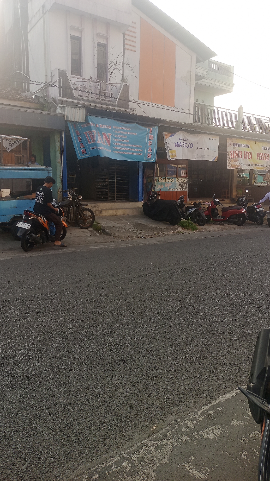 The image shows a street view in front of several shops. The shops are of a commercial nature, one with a blue banner displaying text that appears to be in Indonesian. There are several parked motorcycles and a blue truck. A young man is sitting on a motorcycle in the foreground. The street is paved, and there is a sidewalk in front of the shops. The buildings are two stories high with an orange facade on the upper level of one building. The scene appears to be in a town or city, likely in Indonesia given the text on the banner and the architecture. The time of day appears to be daytime, possibly late afternoon due to the lighting conditions. There are no obvious signs of distress or unusual activity. The overall impression is of a typical street scene in a residential or commercial area.