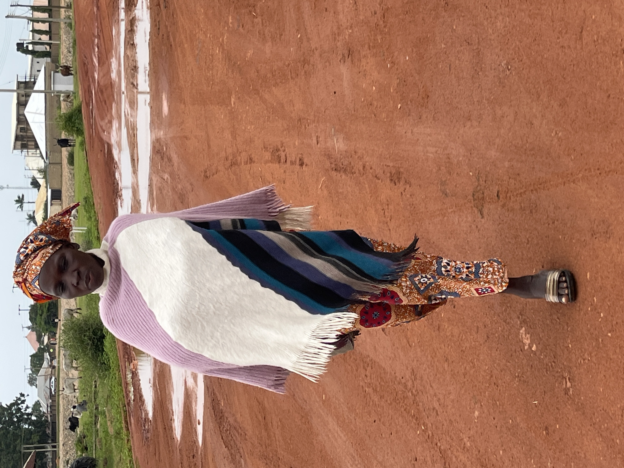 In Bukuru, Nigeria, a woman walks on a dirt road after a rain, with puddles reflecting the overcast sky. She wears a brightly colored headwrap, a striped poncho over a patterned tunic, and sandals. In the background, cattle graze and buildings under construction dot the landscape.